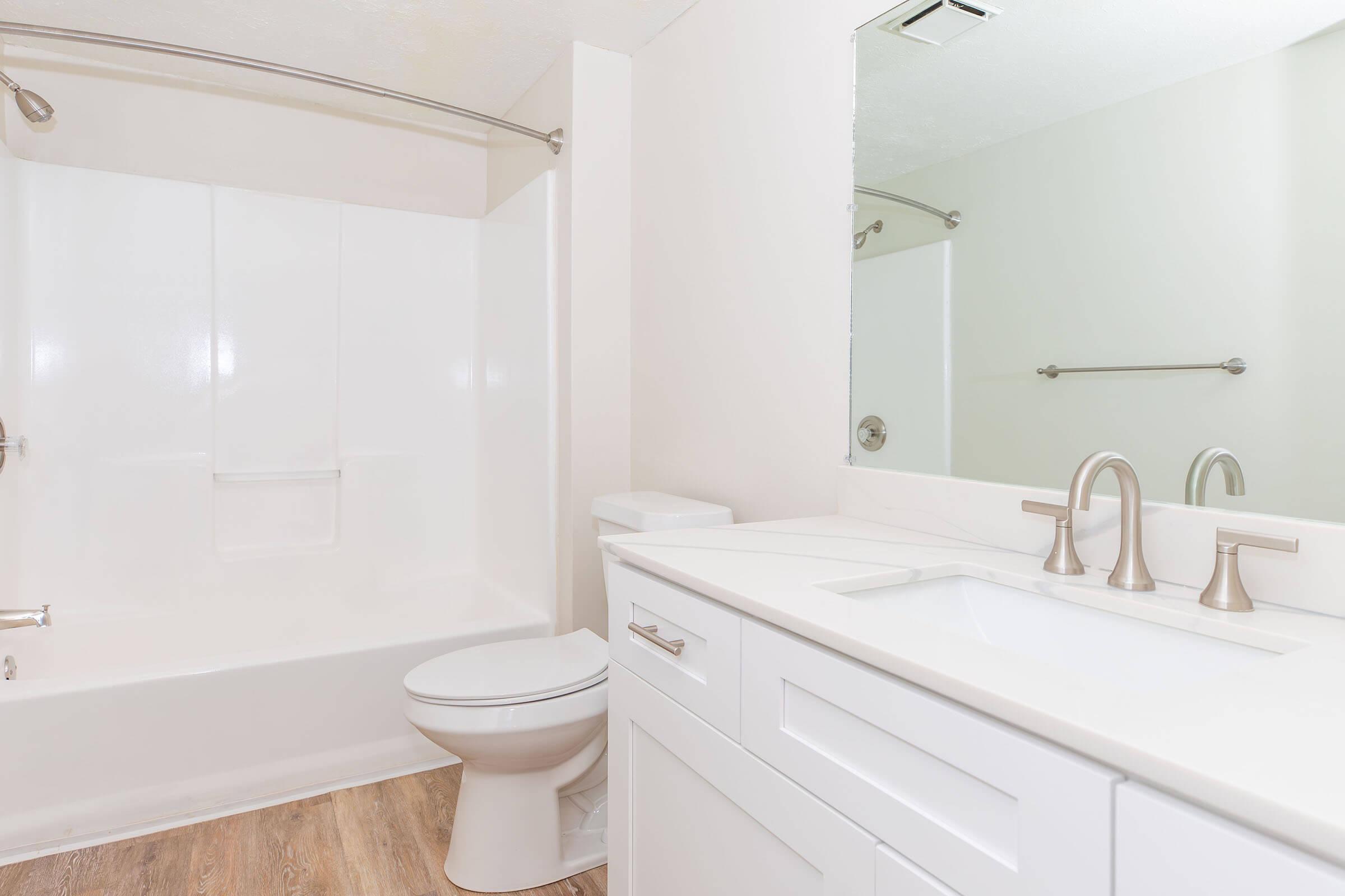A modern bathroom featuring a white bathtub with a shower, a toilet, and a sleek vanity with a sink. The walls are painted light colors, and there is a large mirror above the sink. The flooring is a light wood finish, creating a clean and bright atmosphere.