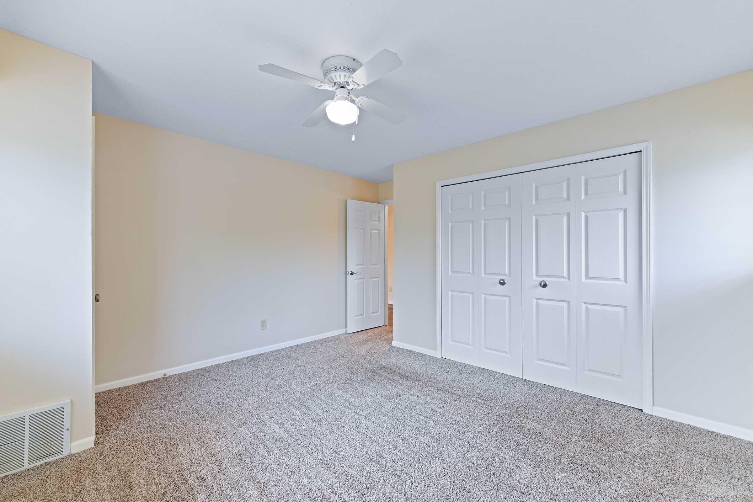 Empty room featuring light-colored walls and carpeted flooring. A ceiling fan is mounted in the center, and there are two closet doors on one wall. A door leading to another room is visible in the background. The overall ambiance is bright and inviting, ideal for furnishing and personalization.