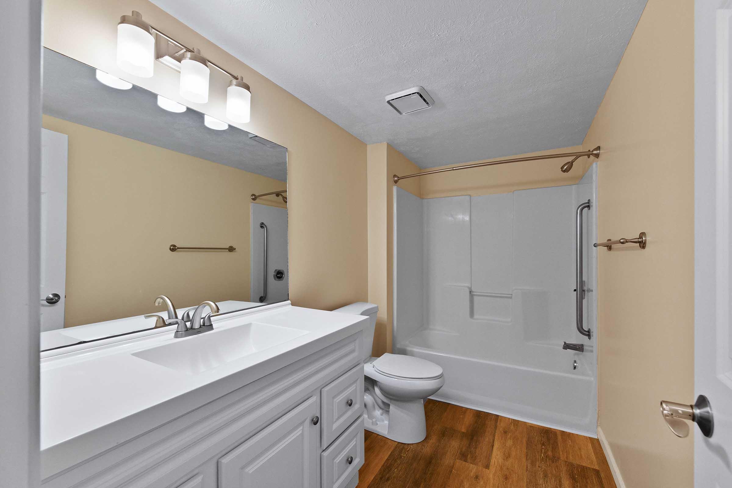 A modern bathroom featuring a white vanity with a large mirror, three light fixtures above, a toilet, and a bathtub with shower. The walls are painted a soft beige, and the flooring is a wood-like material. The overall design is clean and contemporary.