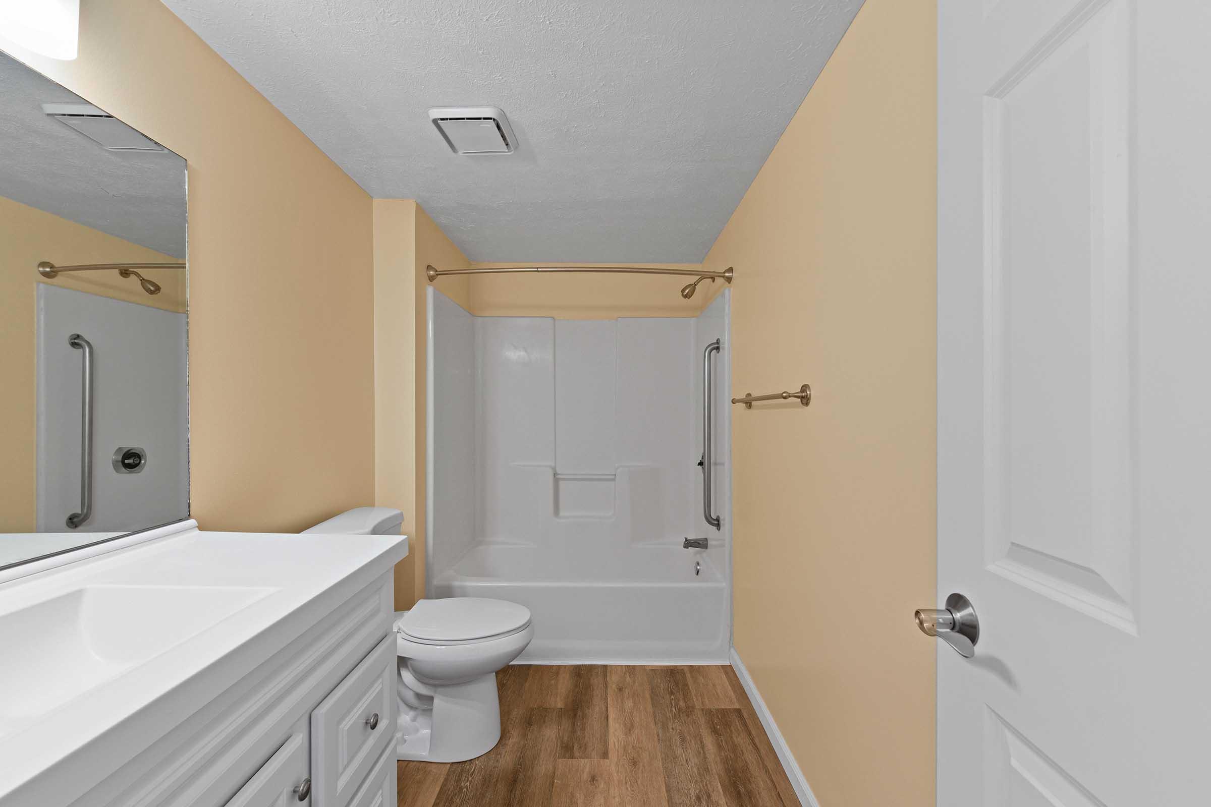 A clean and modern bathroom featuring a white vanity with a mirror, a bathtub with a shower rod, a toilet, and light brown wooden flooring. The walls are painted a soft yellow, creating a bright and inviting atmosphere.