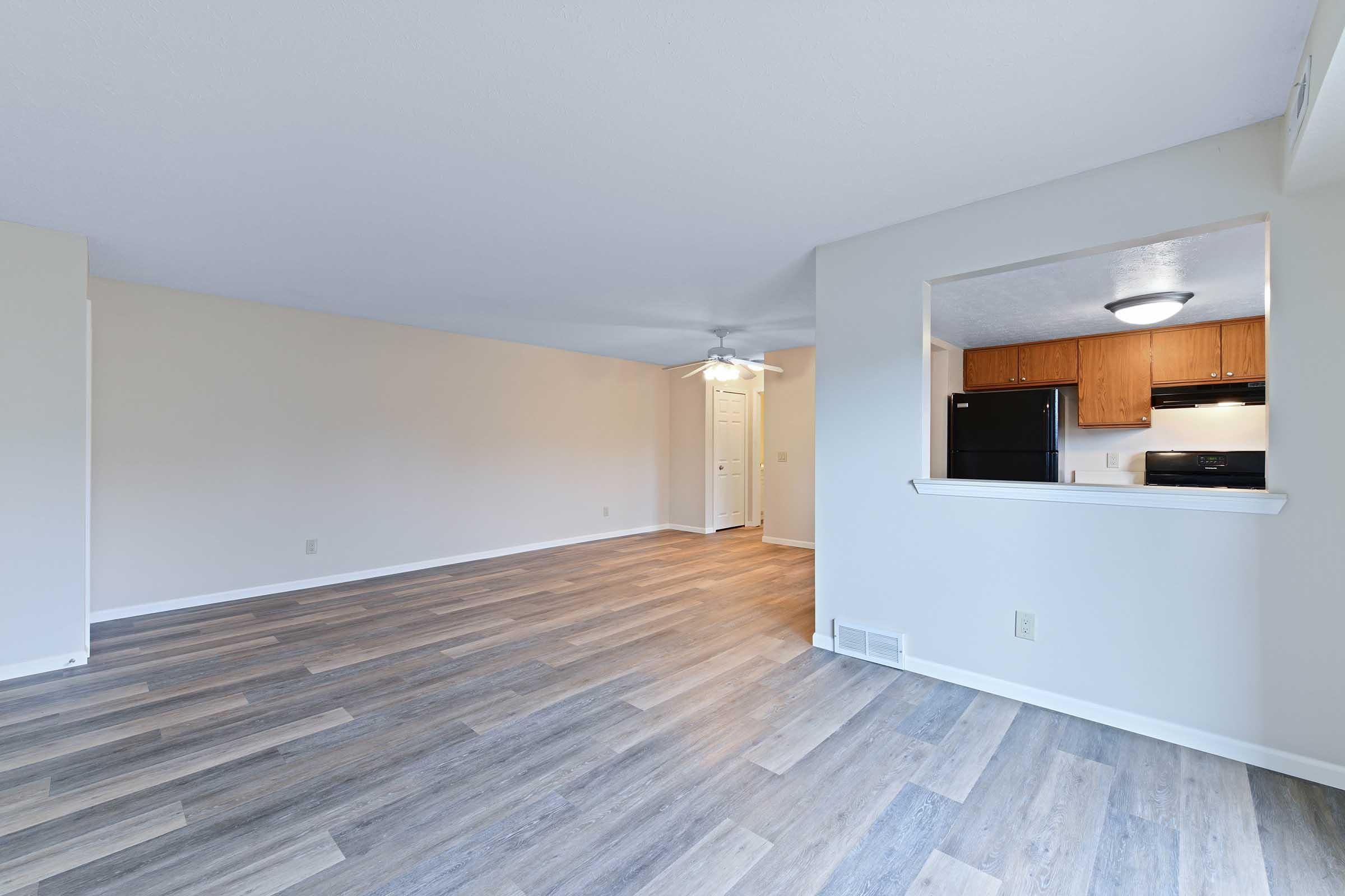 A spacious, unfurnished living area with light-colored walls and a ceiling fan. The flooring features light wood-like planks. A pass-through kitchen area is visible, showing wooden cabinets and black appliances. Natural light fills the space, creating an open and airy atmosphere.