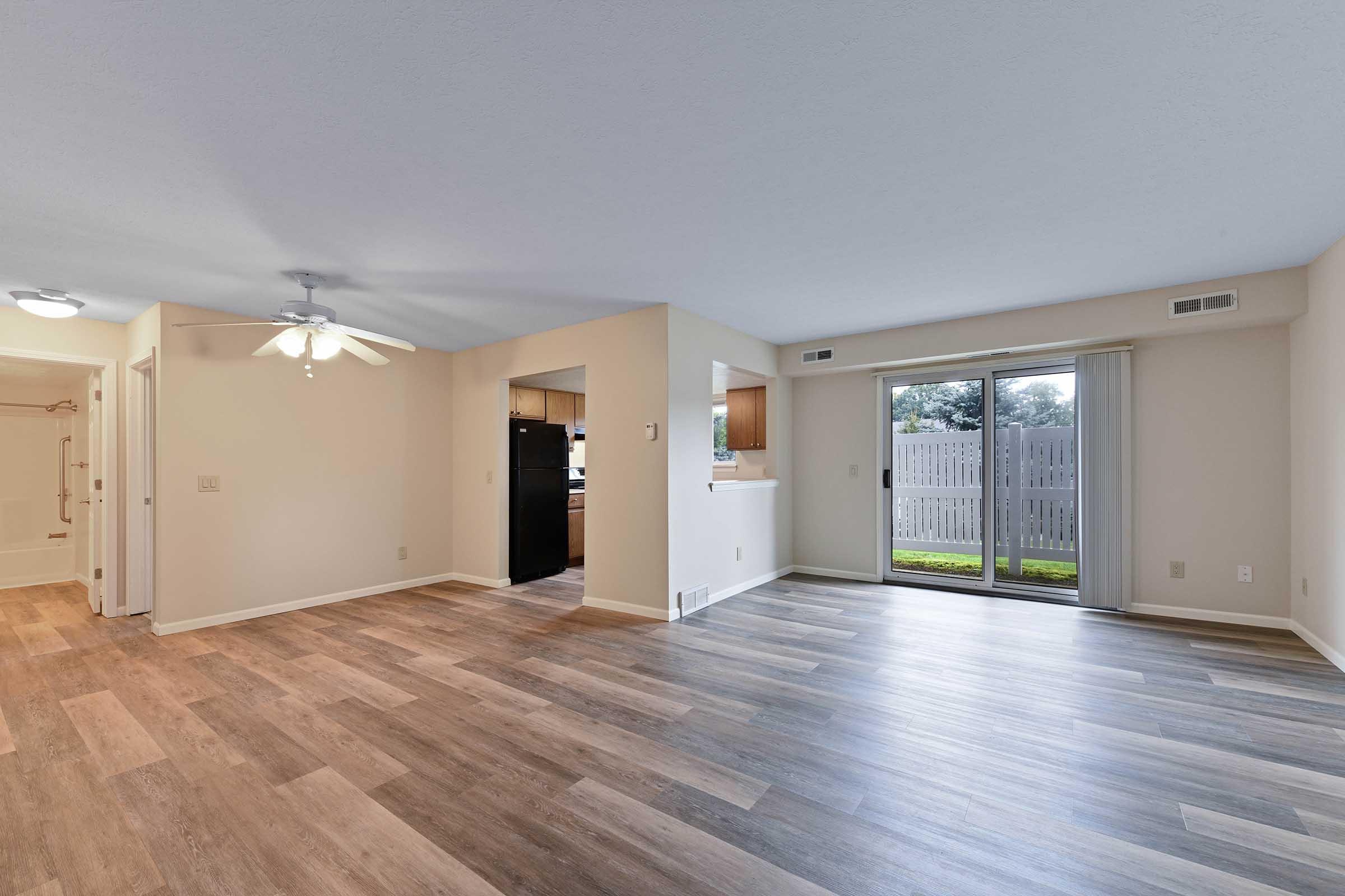 Spacious living area featuring wood-like flooring, ceiling fan, and large sliding glass door leading to an outdoor space. The kitchen is partially visible with dark cabinetry. A bathroom entrance is seen on the left, and natural light brightens the room from multiple windows.