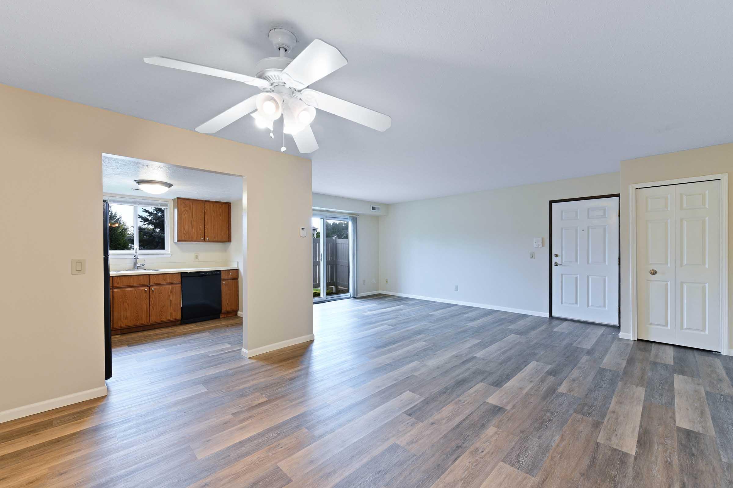 Spacious interior of an apartment featuring a living area with laminate flooring, a ceiling fan, and access to a sliding glass door. Adjacent is a kitchen with wooden cabinets and a window, providing natural light. Two doors lead to separate areas, enhancing the open layout.