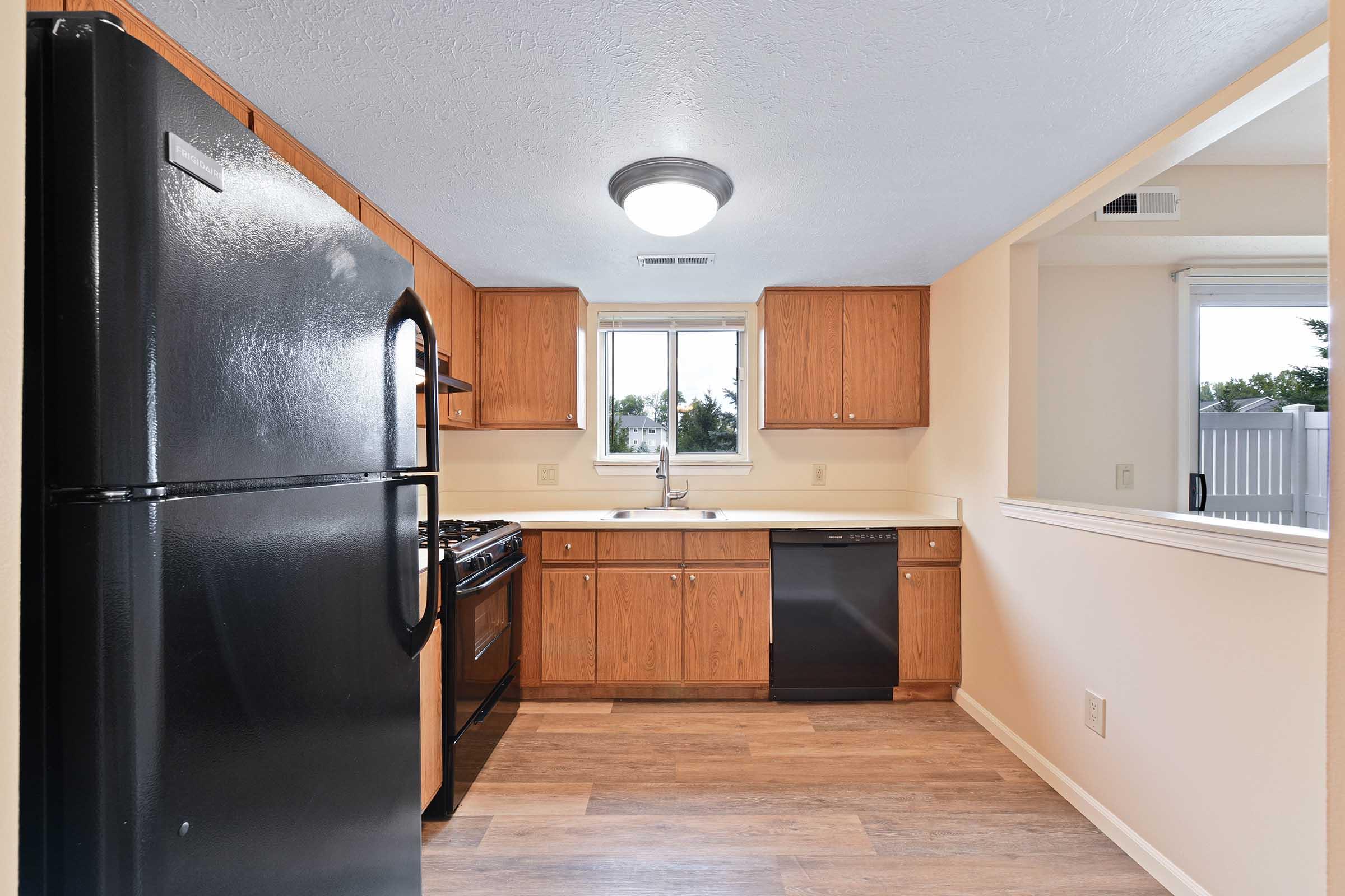 A modern kitchen featuring wooden cabinets, black appliances including a refrigerator and dishwasher, and a gas stove. Natural light fills the space through a window, with light-colored walls and a wood-style floor creating a warm atmosphere.