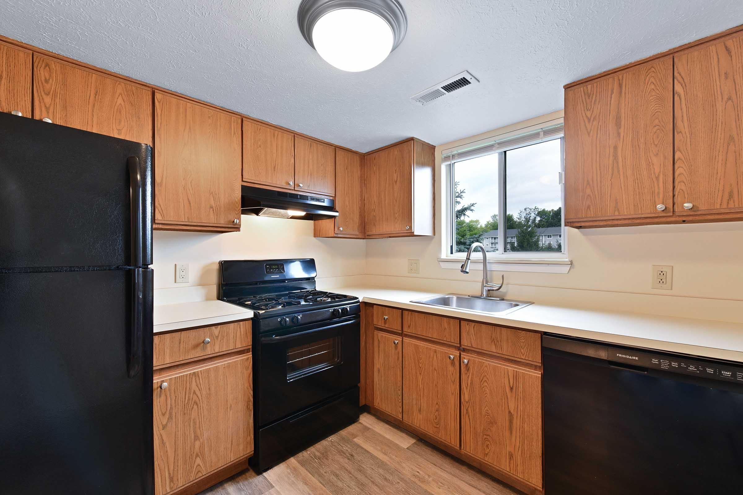 A modern kitchen featuring wooden cabinets, a black refrigerator, a black gas stove, and a sink below a window. The countertops are light-colored, and there is a black dishwasher next to the sink. Natural light brightens the space from the window, creating an inviting atmosphere.