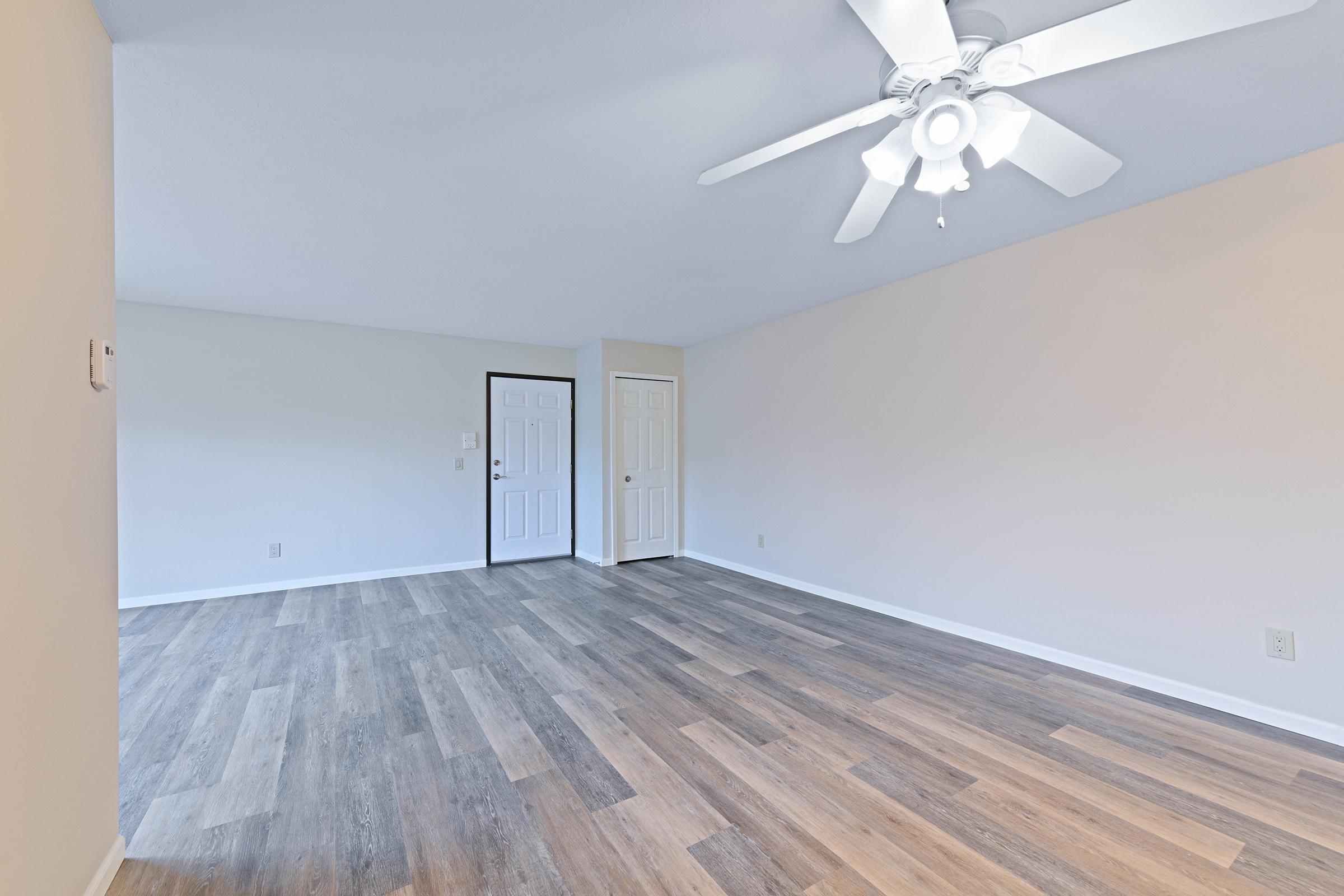 A spacious and empty room featuring light-colored walls, a ceiling fan with white blades, and a combination of light and dark wood-like flooring. Two closed white doors are visible on one side, providing access to adjacent areas. The room is well-lit with natural light, creating an inviting atmosphere.