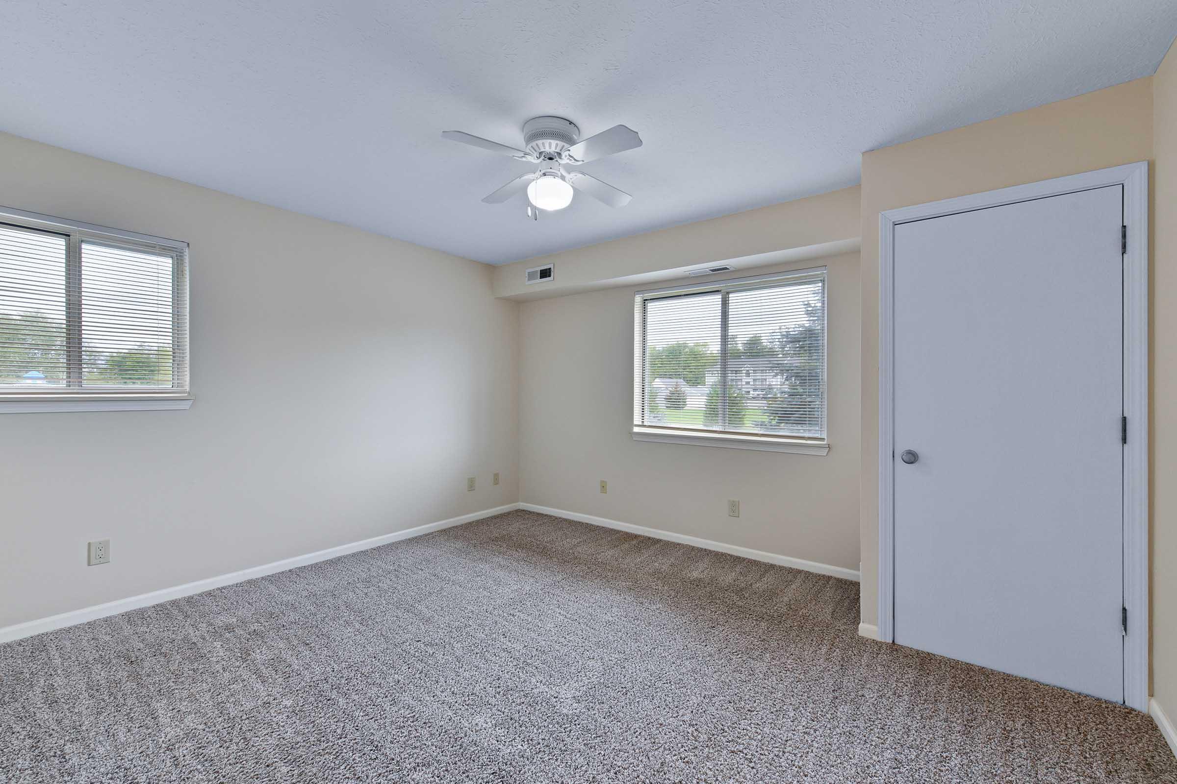 Empty room with light beige walls and a ceiling fan. Two windows with blinds allow natural light, and the floor is covered in light gray carpet. A white door is visible on the right side, and there is minimal furniture, creating a spacious and airy feel.