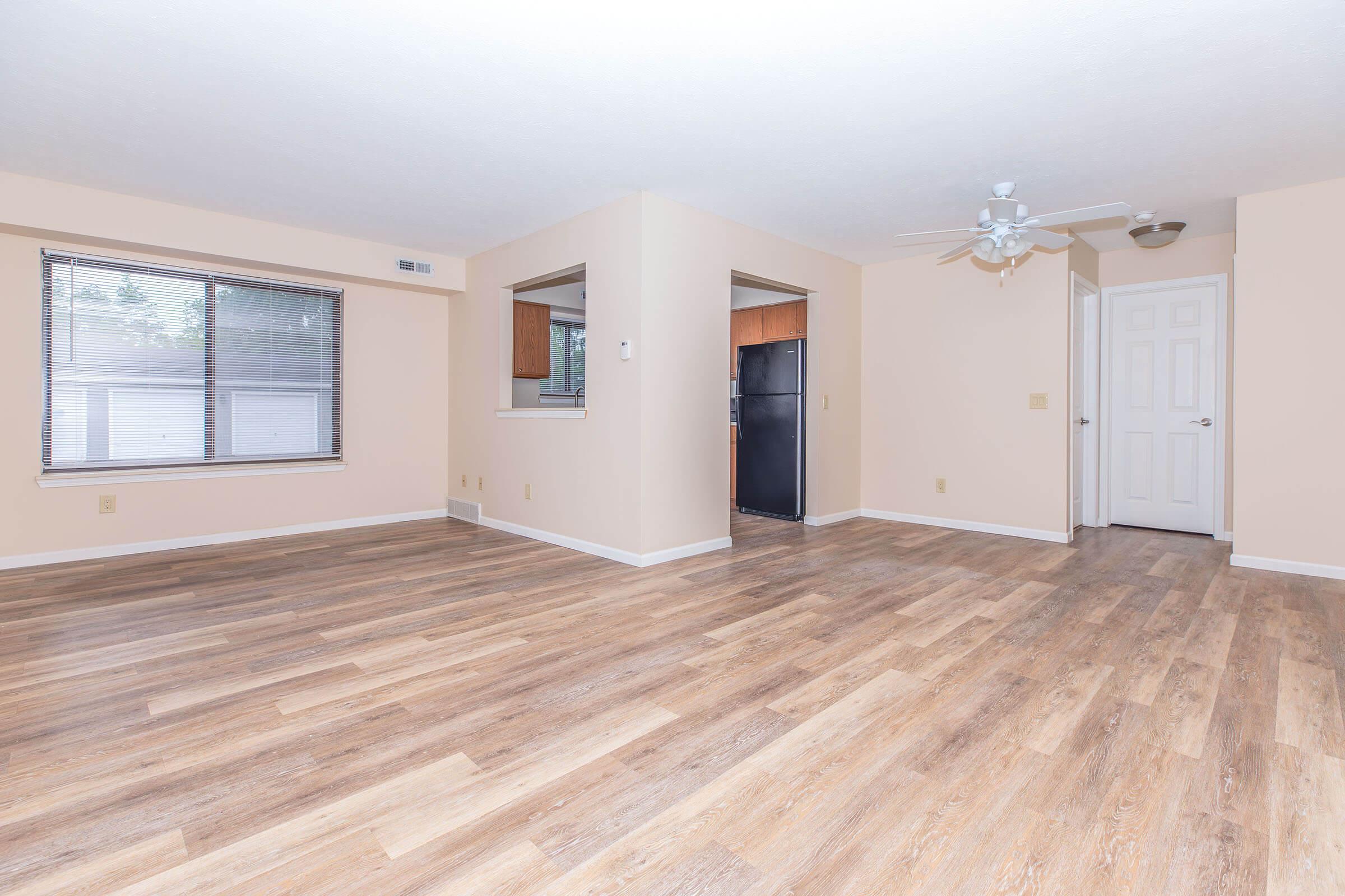 A spacious, empty living room with light beige walls and hardwood-style flooring. A ceiling fan is installed, and there are large windows allowing natural light. In the background, a partial view of a kitchen with a black refrigerator can be seen, along with a doorway leading to another room.