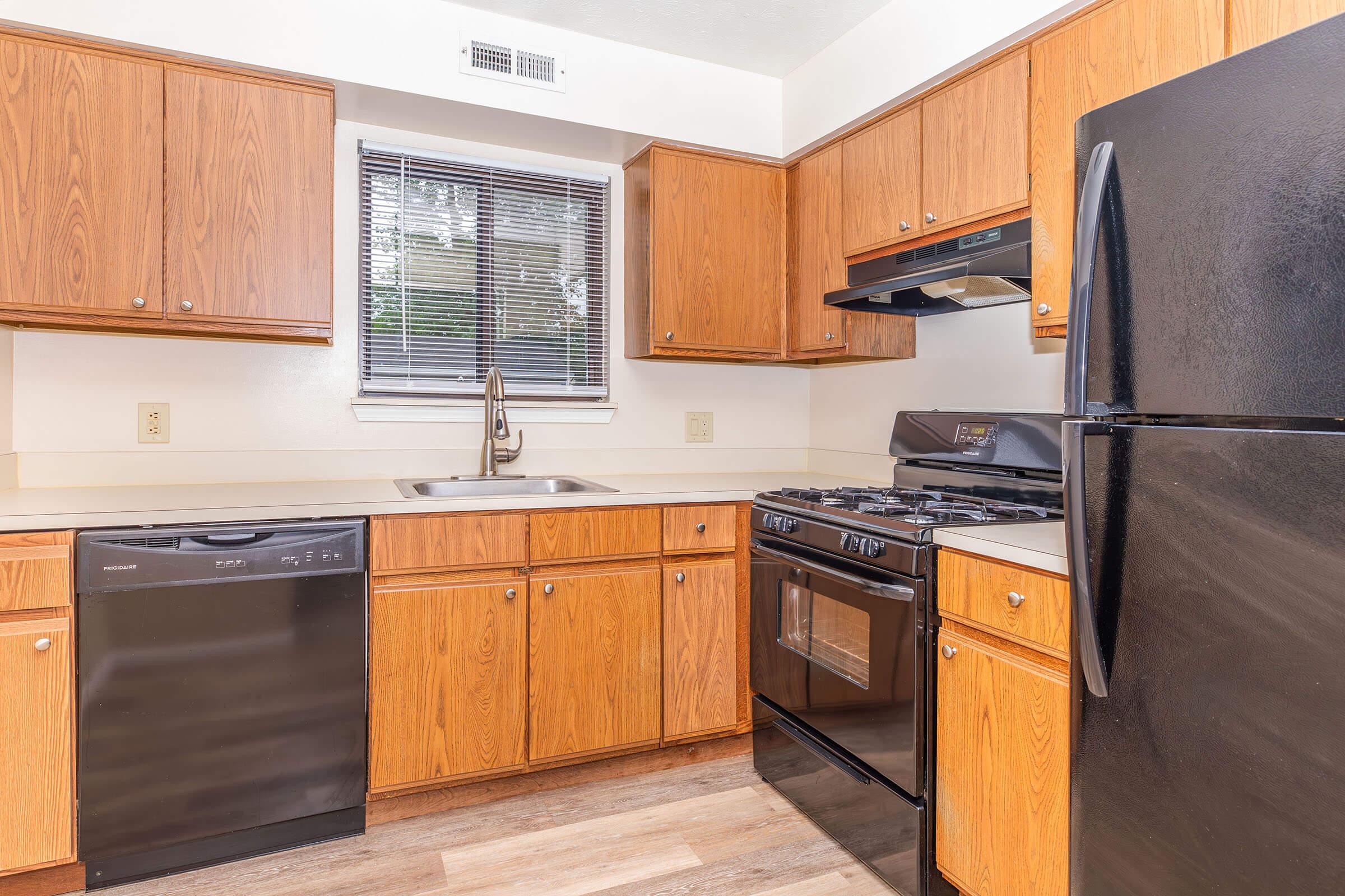 Modern kitchen featuring wooden cabinets, a black refrigerator, a dishwasher, a gas stove, and a stainless steel sink. The countertop is light-colored, and there's a window providing natural light. The flooring is a light wood laminate, creating a warm and inviting atmosphere.