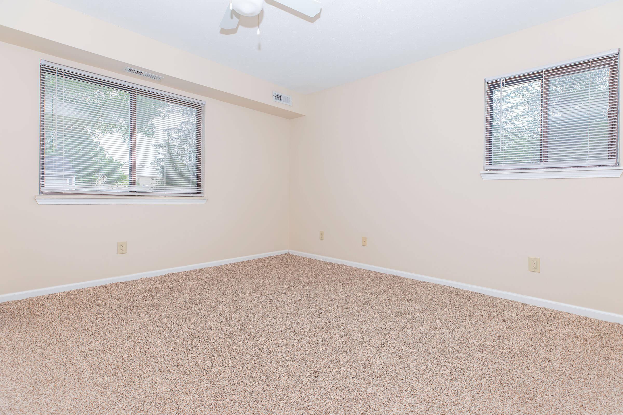 Empty room with light beige walls, two windows with blinds, and a ceiling fan. The floor is covered in beige carpet, creating a spacious and airy atmosphere. No furniture or decorations are present, emphasizing the room's openness.