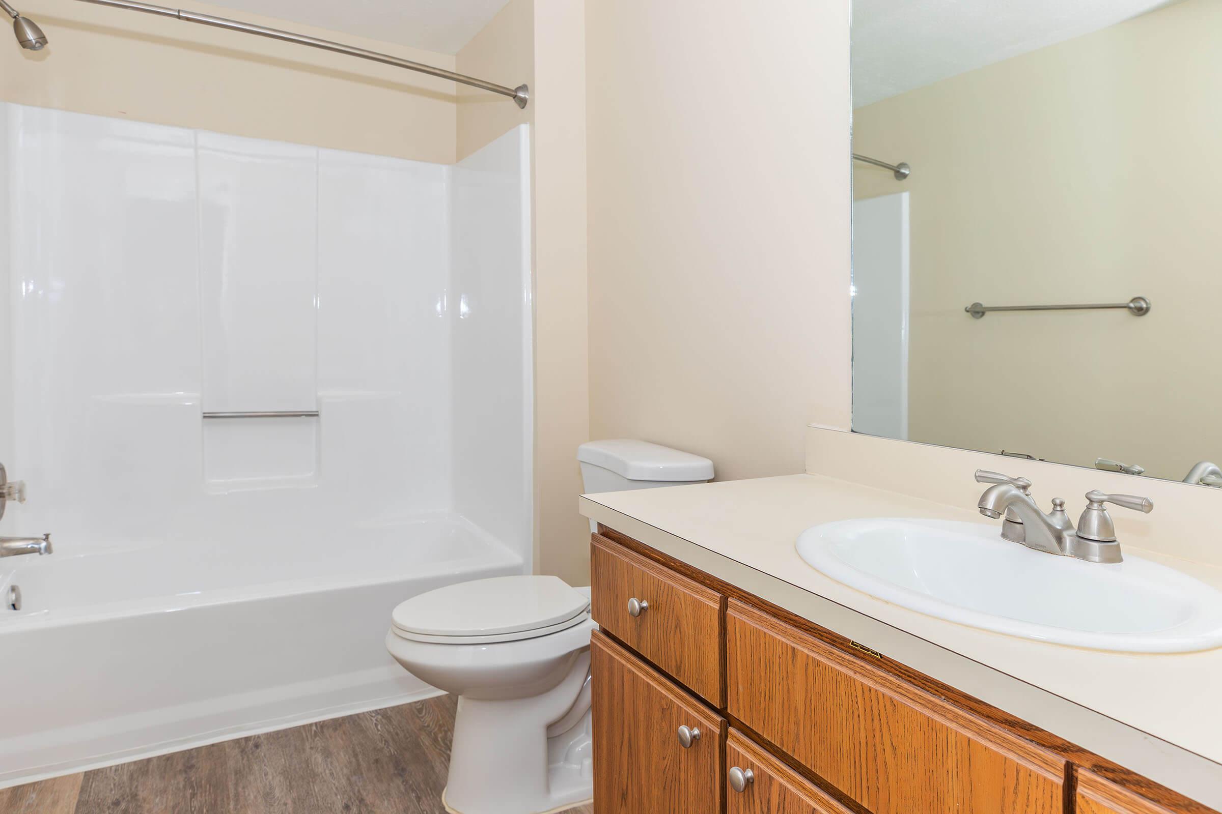 A clean and modern bathroom featuring a white bathtub with a shower, a toilet, and a sink with a mirror above it. The cabinetry is made of light wood, and the walls are painted a soft beige, creating a bright and airy atmosphere.