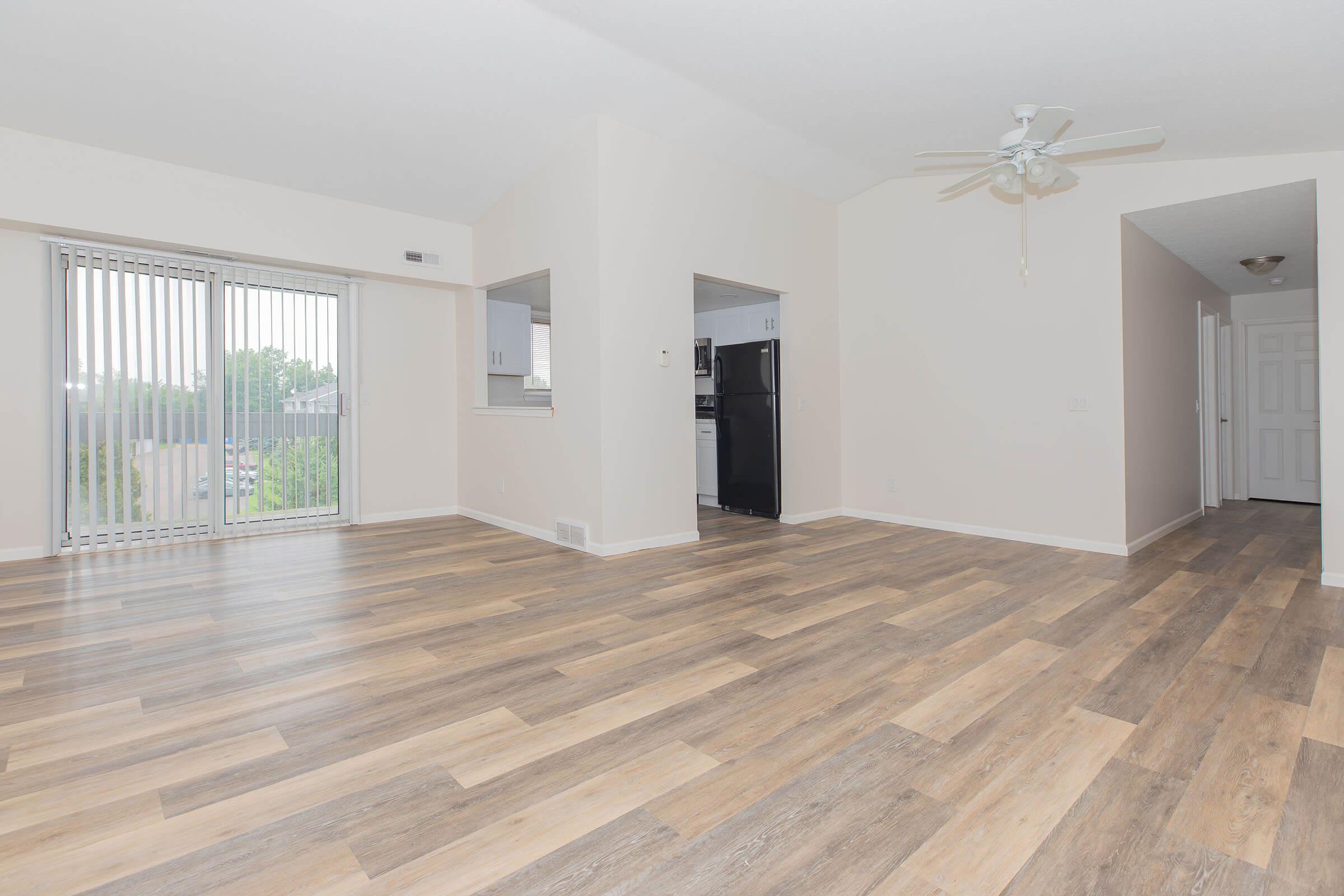 Spacious interior of a modern living room featuring light-colored walls and a large window with vertical blinds. The floor is covered with brown and beige laminate wood. A doorway leads to a kitchen area, and another doorway is visible in the background, suggesting access to other rooms. Bright and airy atmosphere.