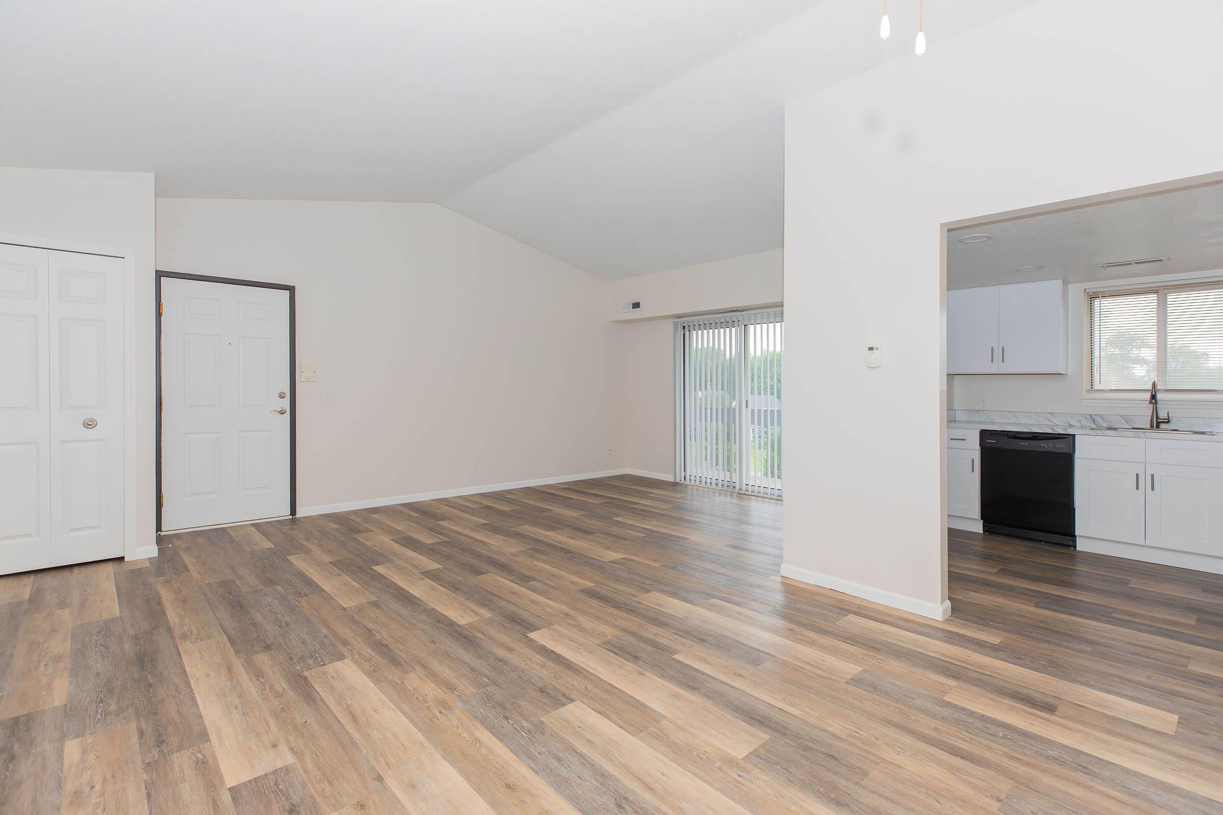 Spacious interior of a modern apartment with light-colored walls and wooden flooring. The room features a tall ceiling, a sliding glass door leading to an outdoor area, and a view into a kitchen with white cabinets and appliances. A closed door and closet are visible on the left.