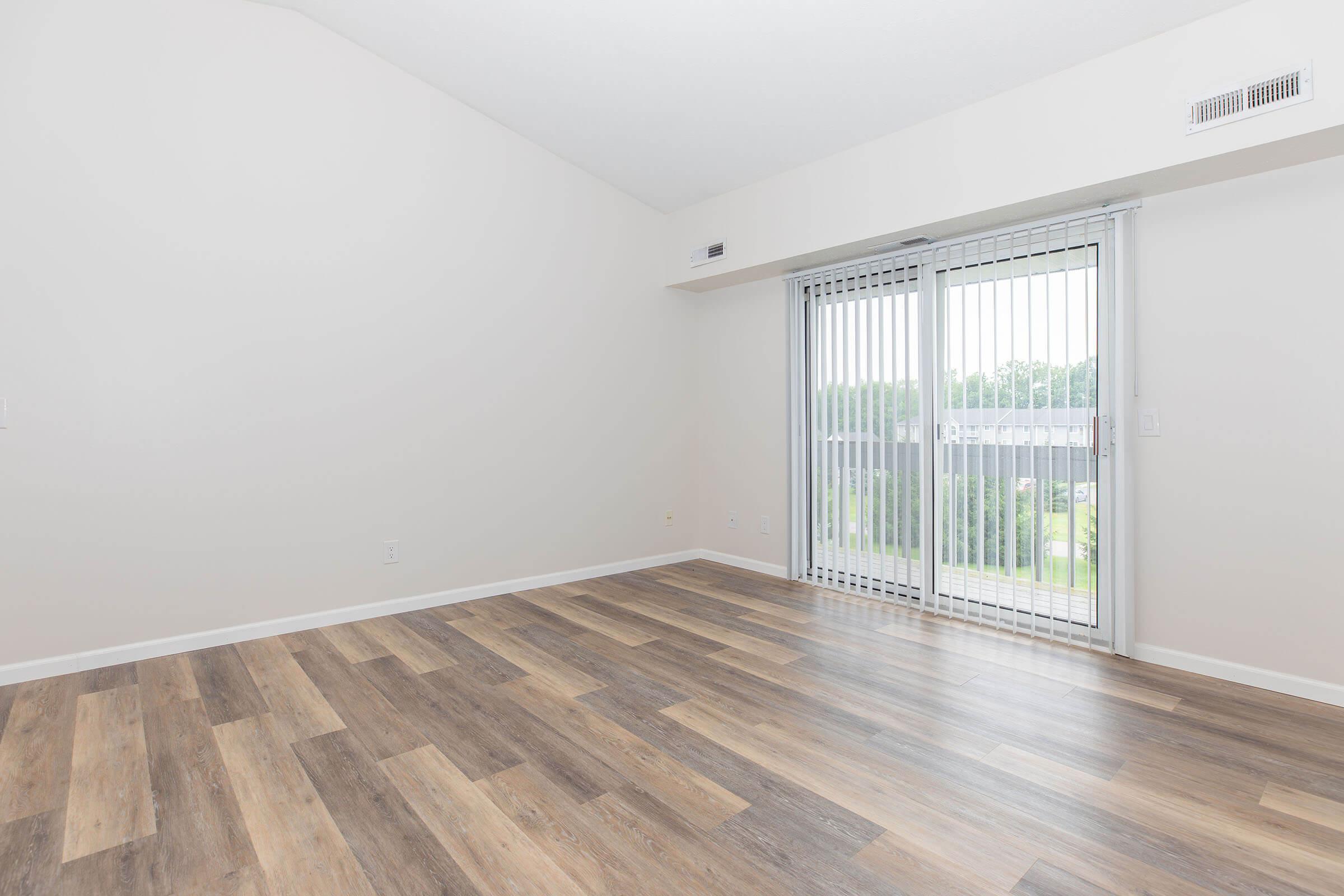 Empty room with light-colored walls and a large sliding glass door leading to an outdoor area. The floor features a mix of light and dark wood-like tiles, providing a modern look. Natural light is streaming in through the door, creating a bright atmosphere.