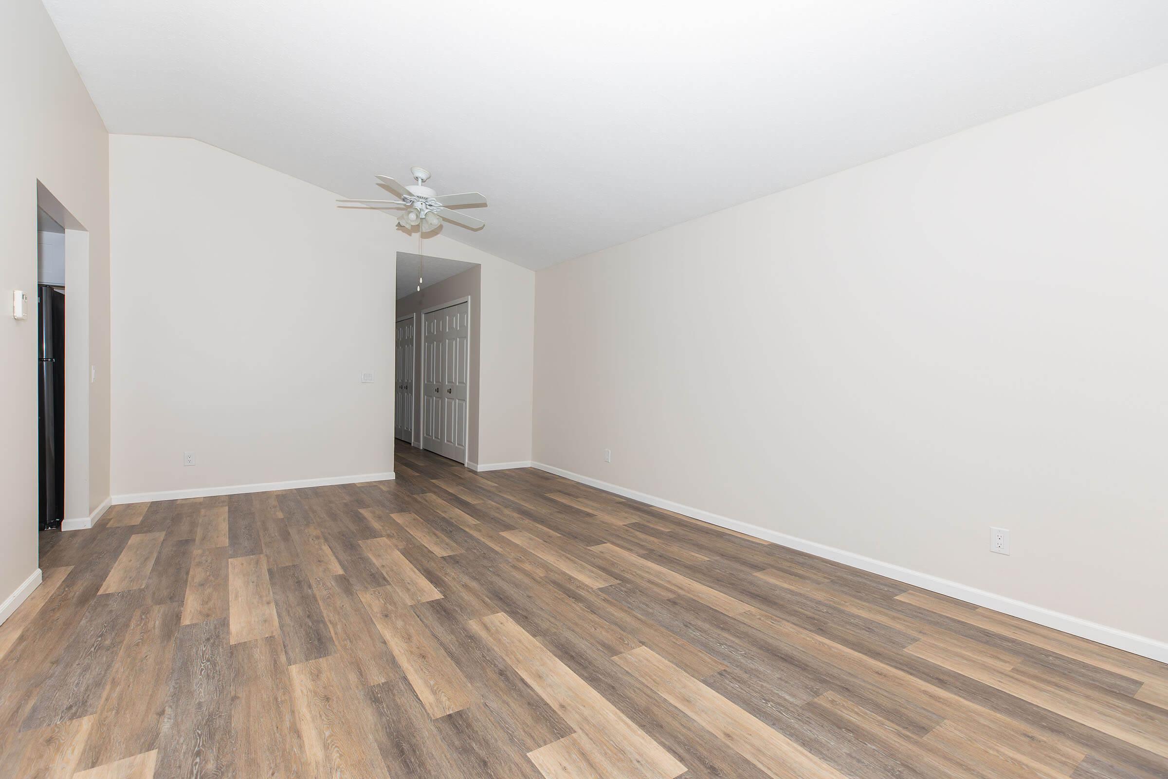 Spacious room with wooden flooring, neutral-colored walls, and a ceiling fan. A doorway leads to another area, and there are multiple doors along one wall, indicating potential bedrooms or storage. The overall atmosphere is open and airy, with plenty of natural light.