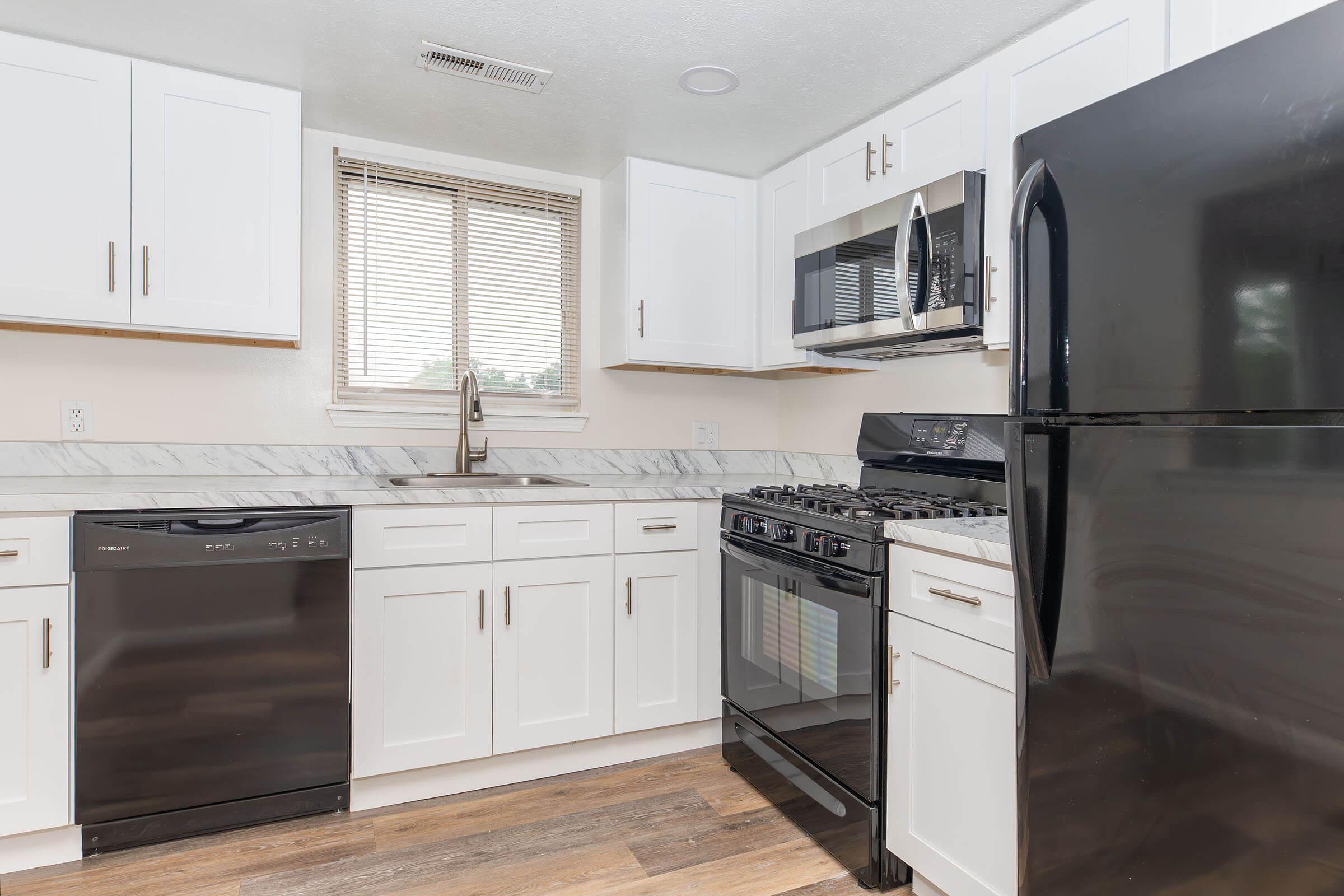 A modern kitchen featuring white cabinetry, a black refrigerator, a stainless steel microwave, and a gas stove. The countertops are marble with a sink below a window. A dishwasher is also present next to the sink, and the flooring is wood-like laminate. Bright and well-lit space.