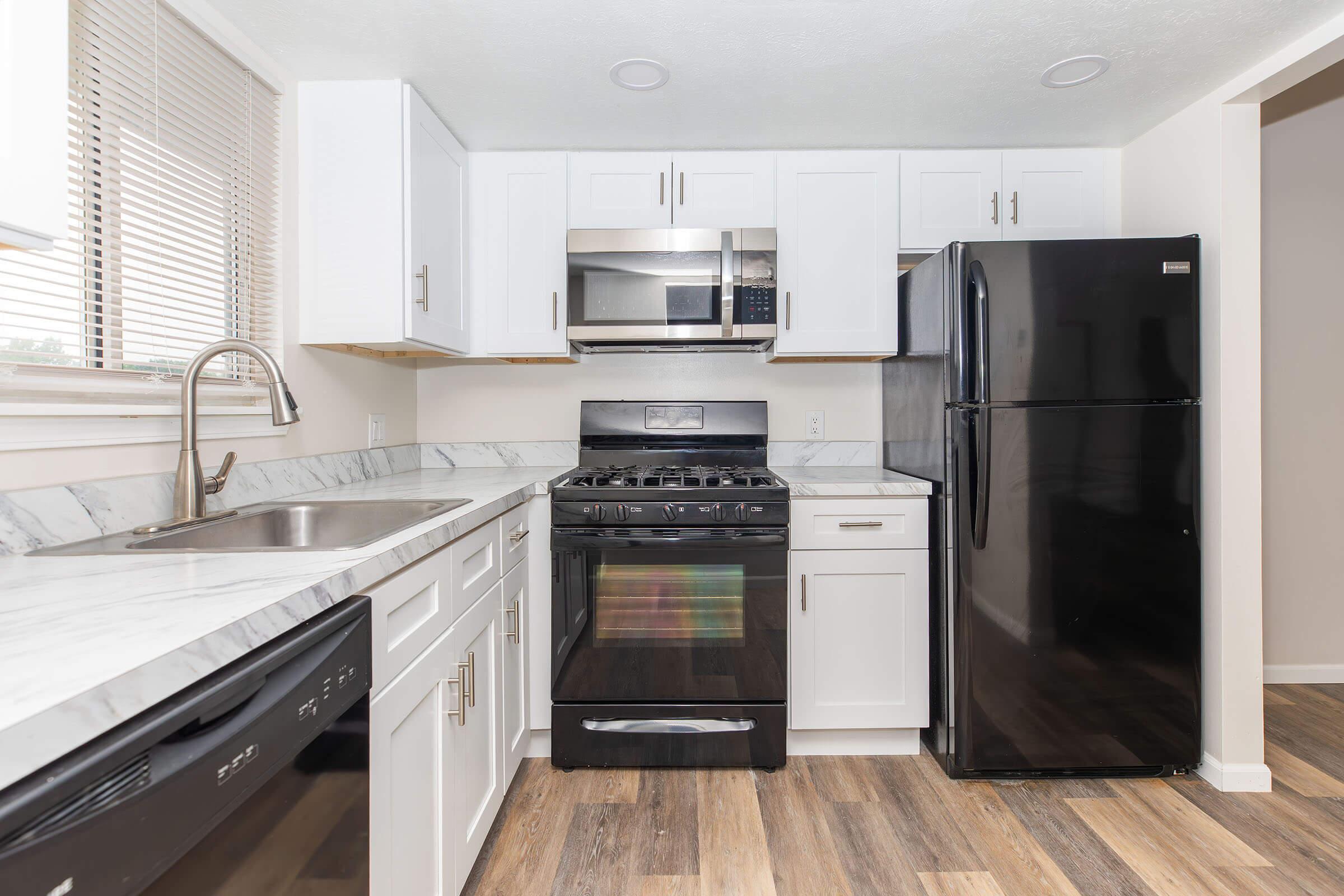 A modern kitchen featuring white cabinets, a stainless steel microwave and stove, a black refrigerator, and a dishwasher. The countertops are marble with a light gray hue. Natural light enters through a window, highlighting the wood-like flooring.