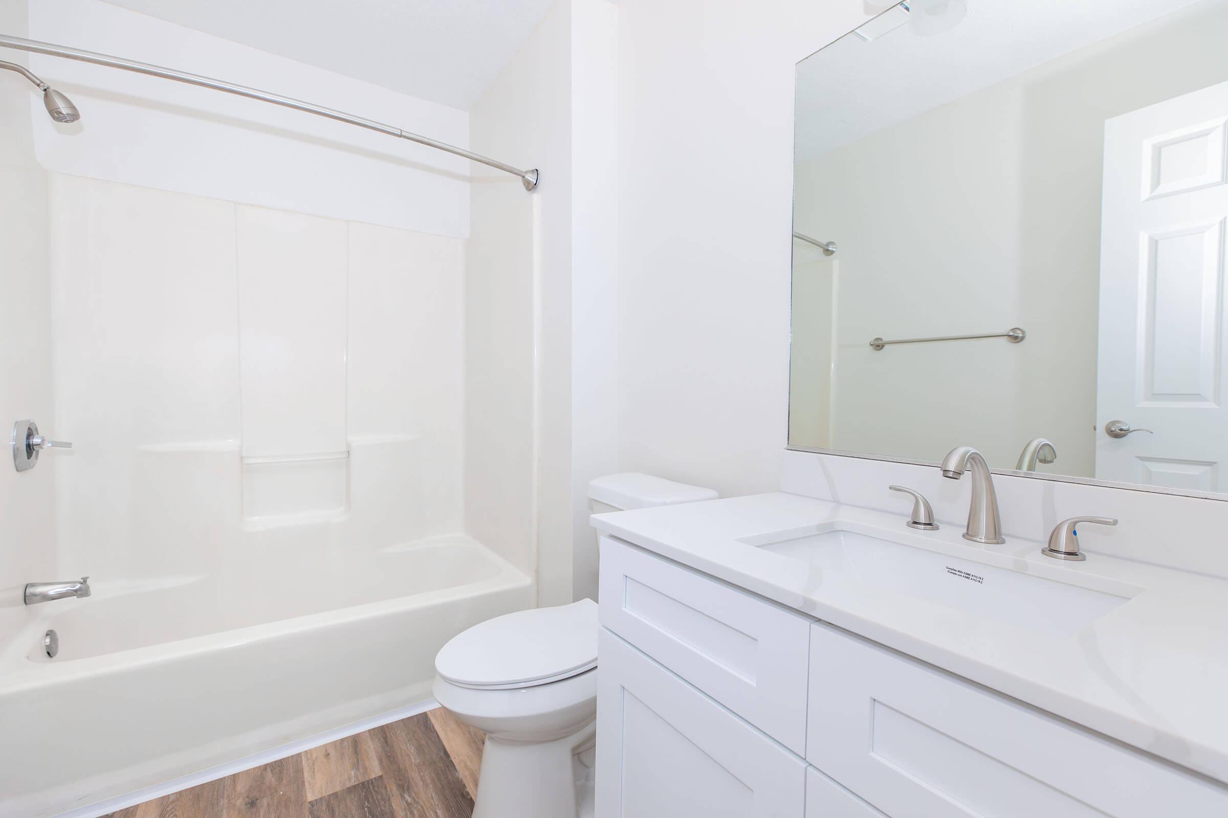 A clean and modern bathroom featuring a white bathtub with a curved shower rod, a white vanity with dual sinks and polished faucets, a toilet, and a large mirror above the sink. The walls are painted in a light color, and the flooring is made of wood-like tiles, contributing to a fresh and bright atmosphere.