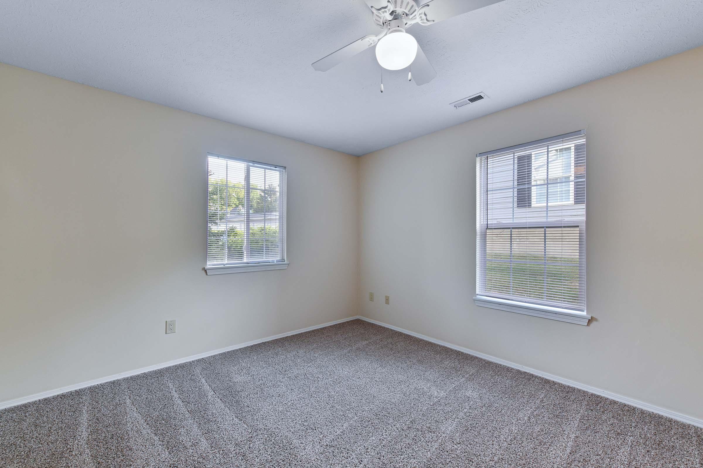 A spacious, empty room with light beige walls and carpet. There are two windows with white blinds allowing natural light to enter. A ceiling fan is mounted on the white ceiling, and there are no furnishings in the room, creating a clean and open atmosphere.