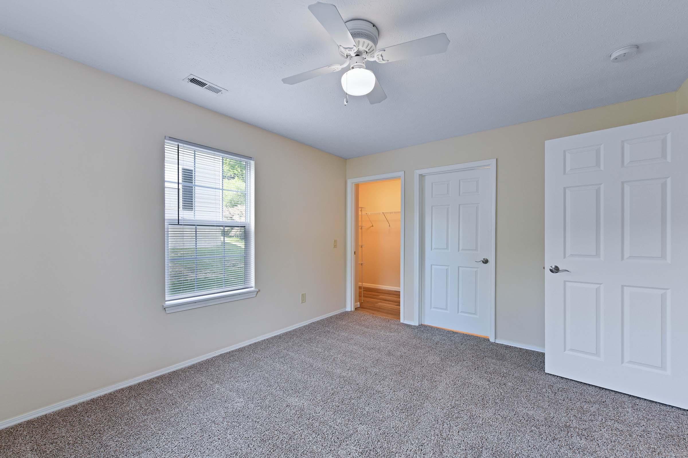 A well-lit, empty bedroom featuring light beige walls and beige carpet. It includes a ceiling fan with a light fixture, a window with window blinds, and two closed doors—one leading to a closet and another leading to a bathroom or hallway.