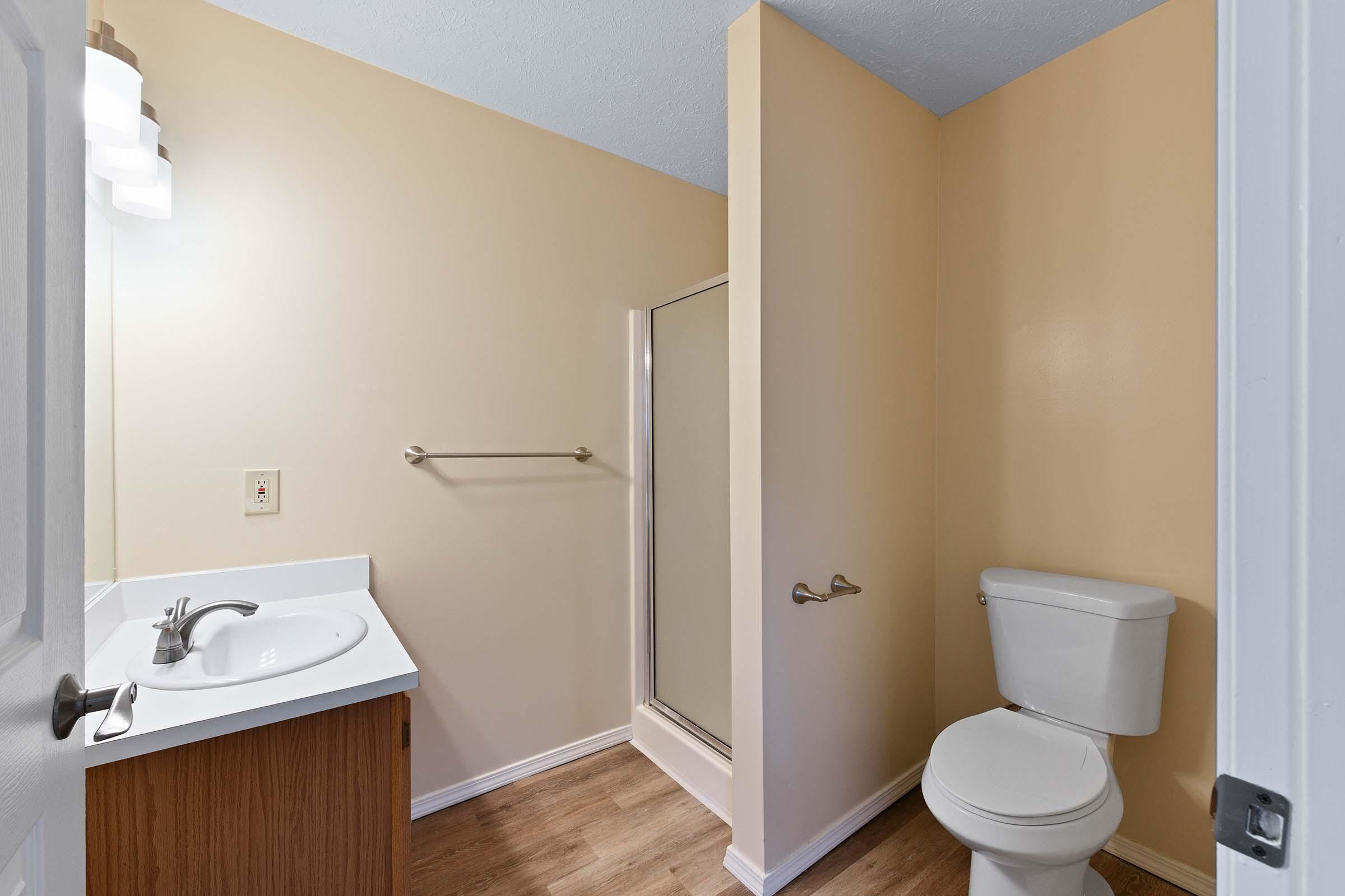 A small bathroom featuring a beige color scheme, a single sink with a mirror above, a toilet, and a corner shower without a door. The flooring is wooden, and a towel rack is mounted on the wall, providing a simple and functional design.
