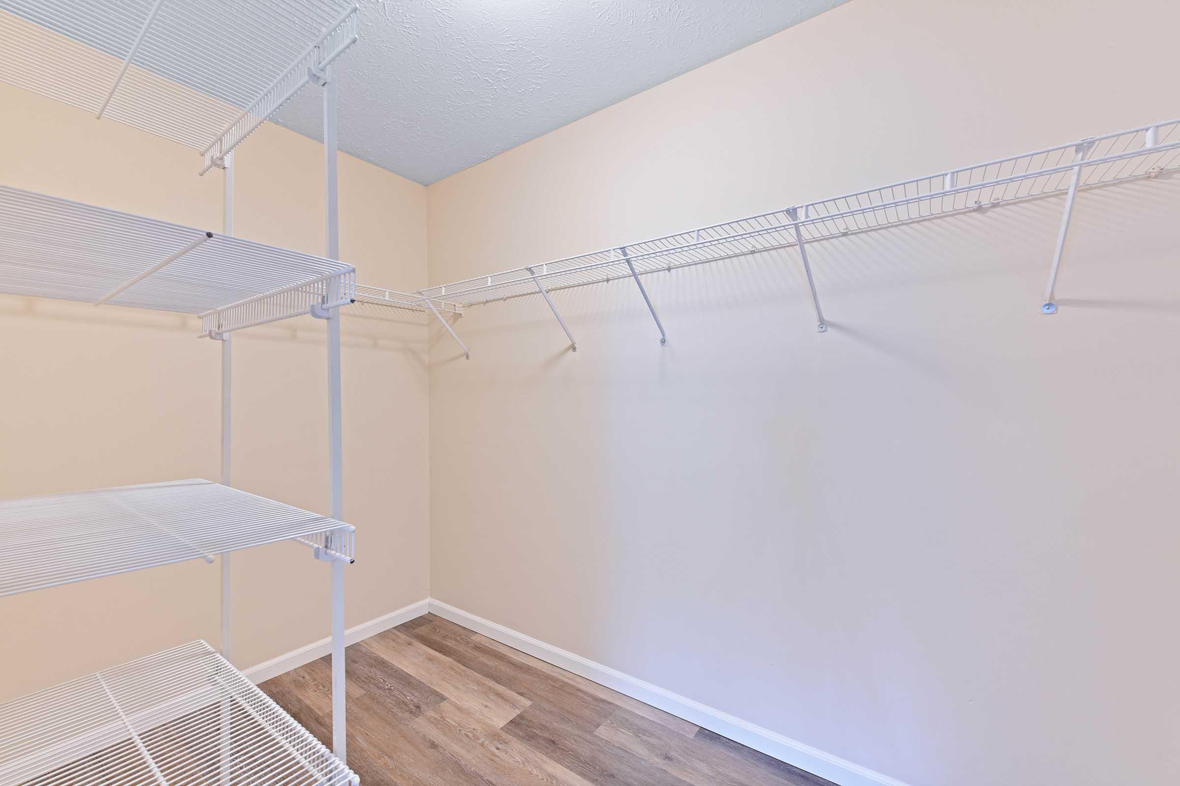 Empty closet with white wire shelving. The walls are painted a light beige, and the floor has a wood-like appearance. The shelving includes several shelves and a hanging rod, providing storage space for various items. The overall atmosphere is clean and simple.