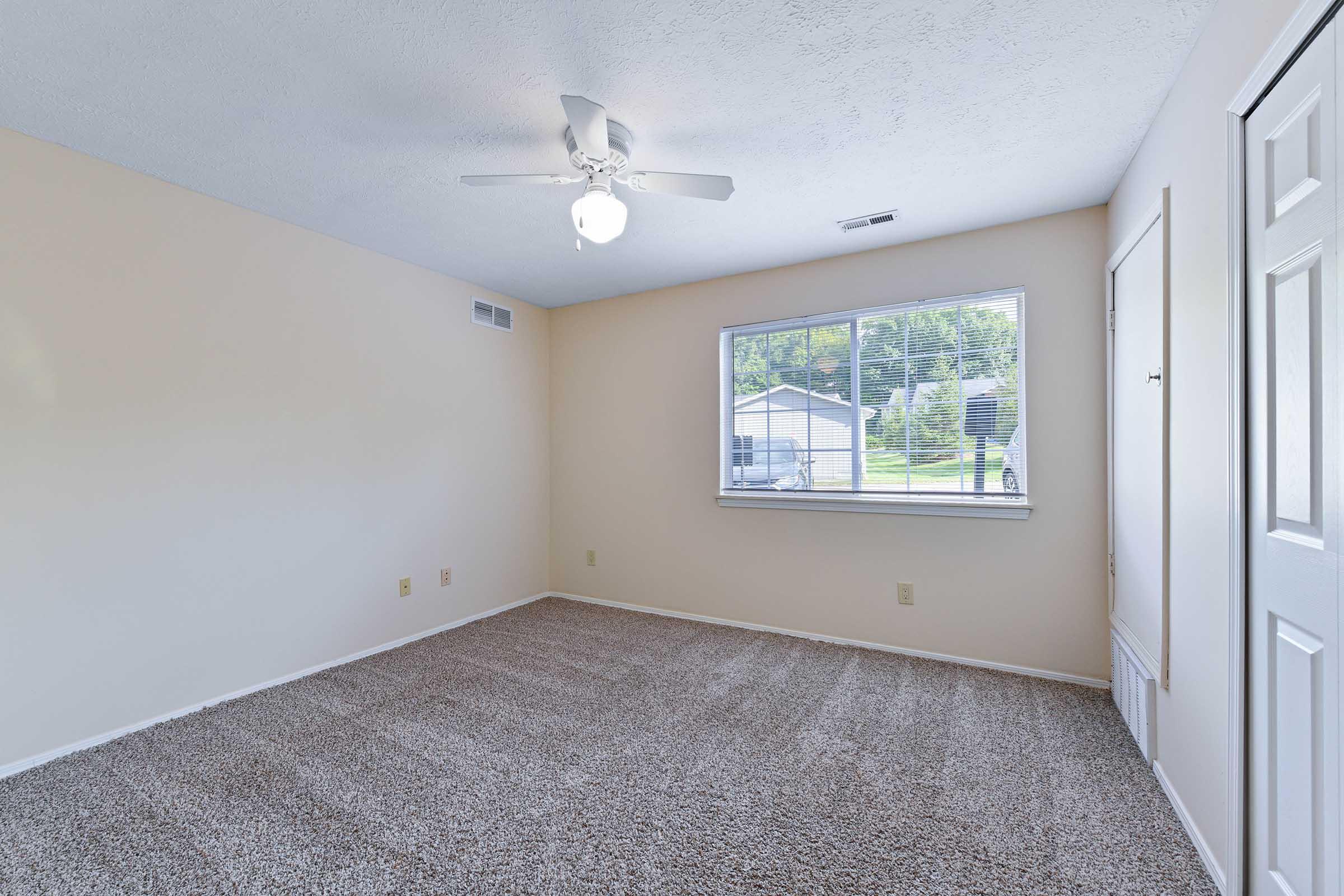 Empty room with light beige walls and plush carpet, featuring a ceiling fan. A large window allows natural light to enter, and there's a closet with sliding doors. View outside shows a glimpse of greenery. The space is clean and ready for furnishing.