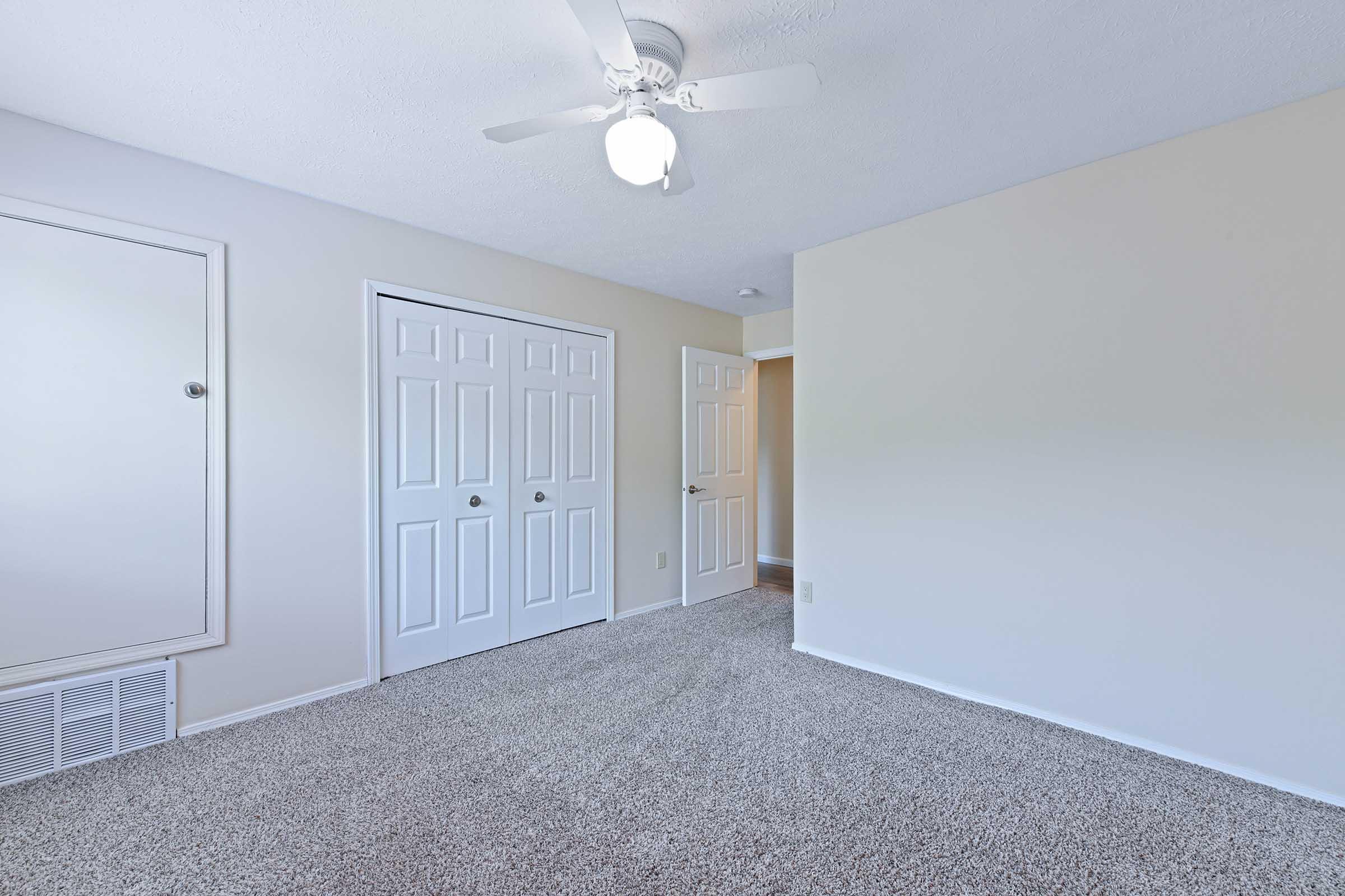 A spacious, empty room with light beige walls and plush gray carpeting. It features a ceiling fan, a mirrored closet door, and white double doors that lead to another area. Natural light brightens the space, creating an airy atmosphere.
