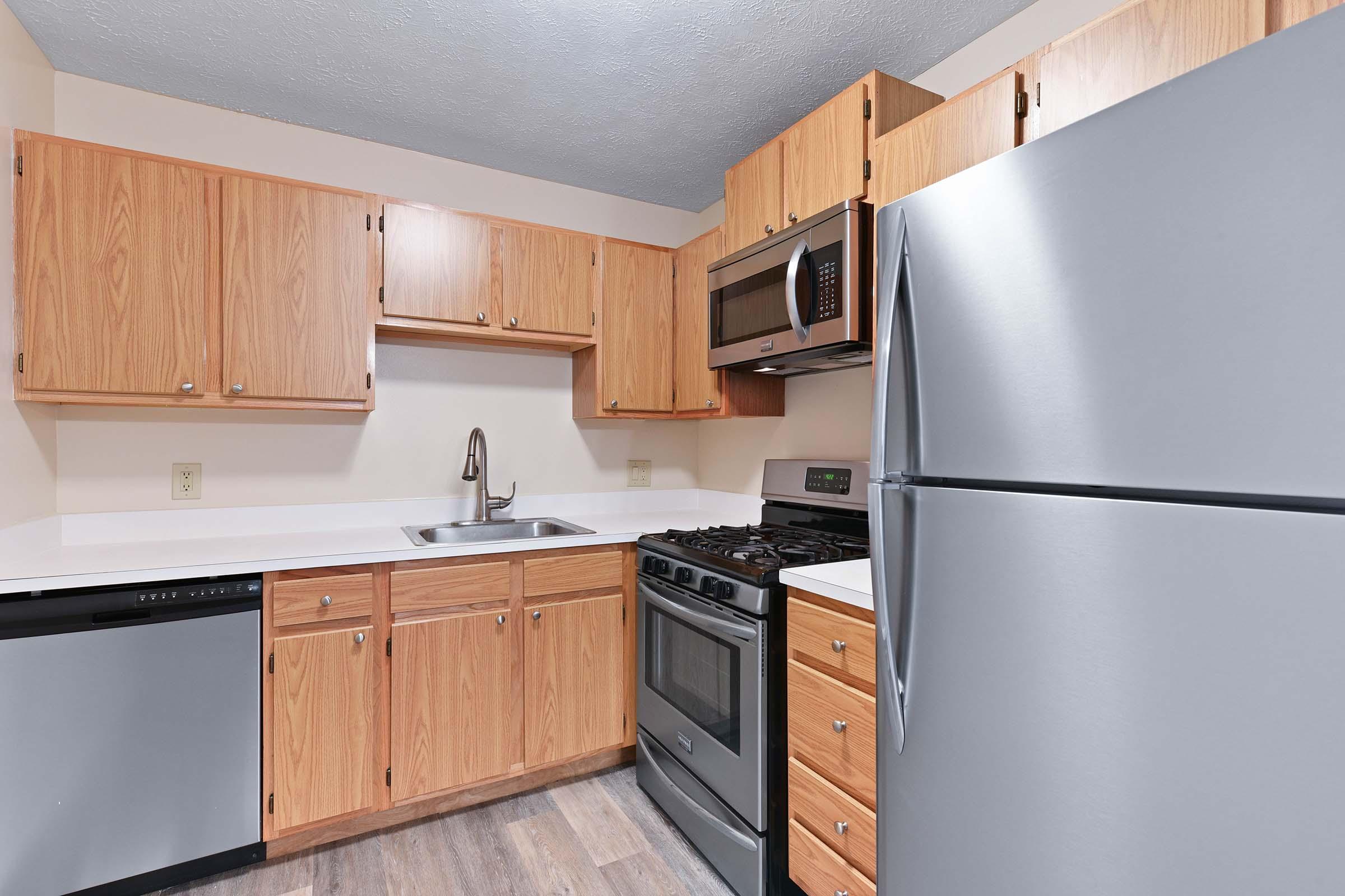 A modern kitchen with light wooden cabinets, stainless steel appliances including a refrigerator, oven, and microwave, a single basin sink, and a dishwasher. The countertop is white, and the floor features a wood-like finish, creating a clean and inviting atmosphere.