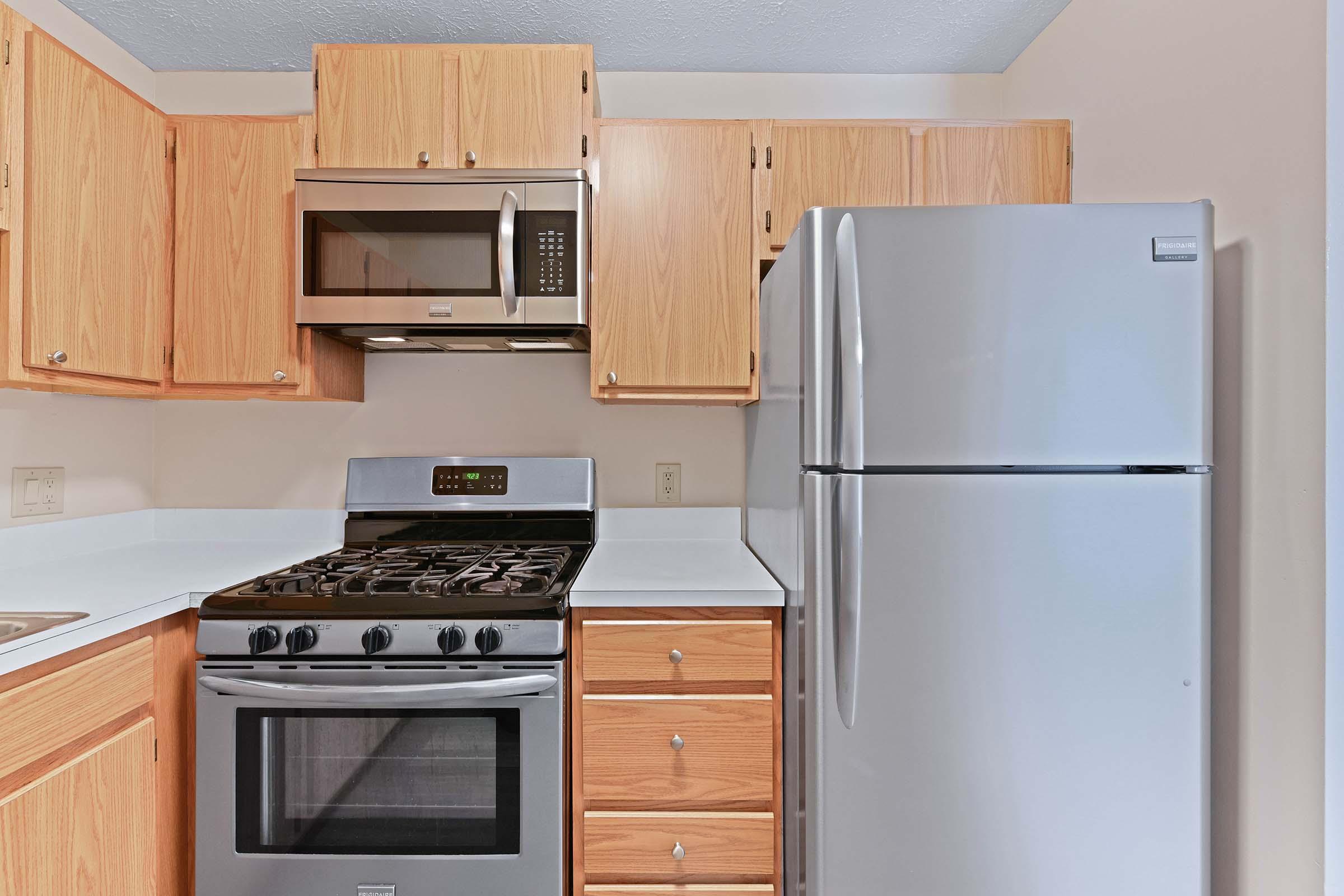 A modern kitchen featuring wooden cabinetry, a stainless steel refrigerator, a microwave, and a gas stove with an oven. The countertops are white, and the overall design is clean and functional, emphasizing a contemporary look.