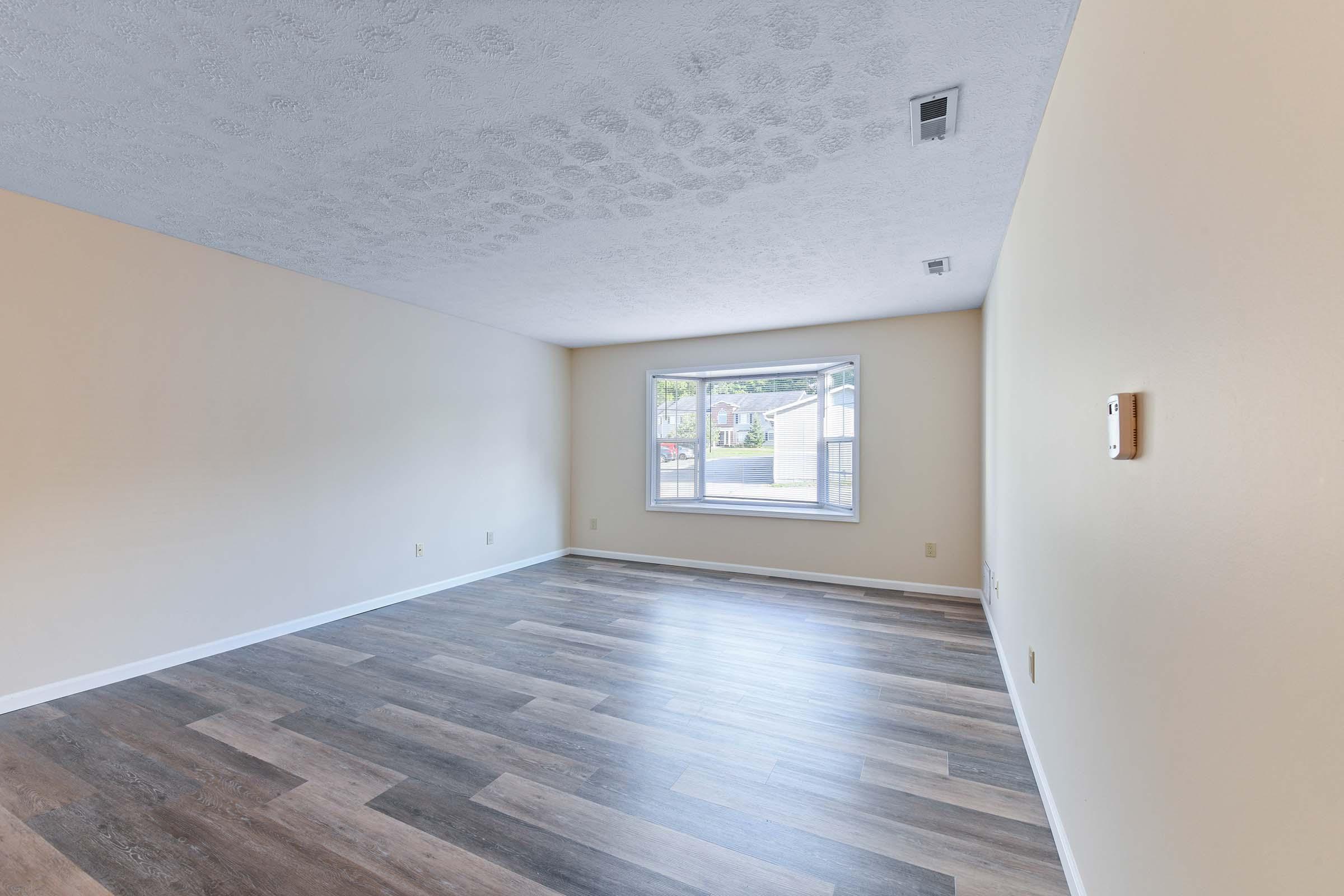 A spacious, empty room with light-colored walls and a large window letting in natural light. The floor features a mix of dark and light wood tones. The ceiling has a textured finish, and there is a single electrical outlet on one wall. The overall atmosphere is bright and inviting, ready for decoration.