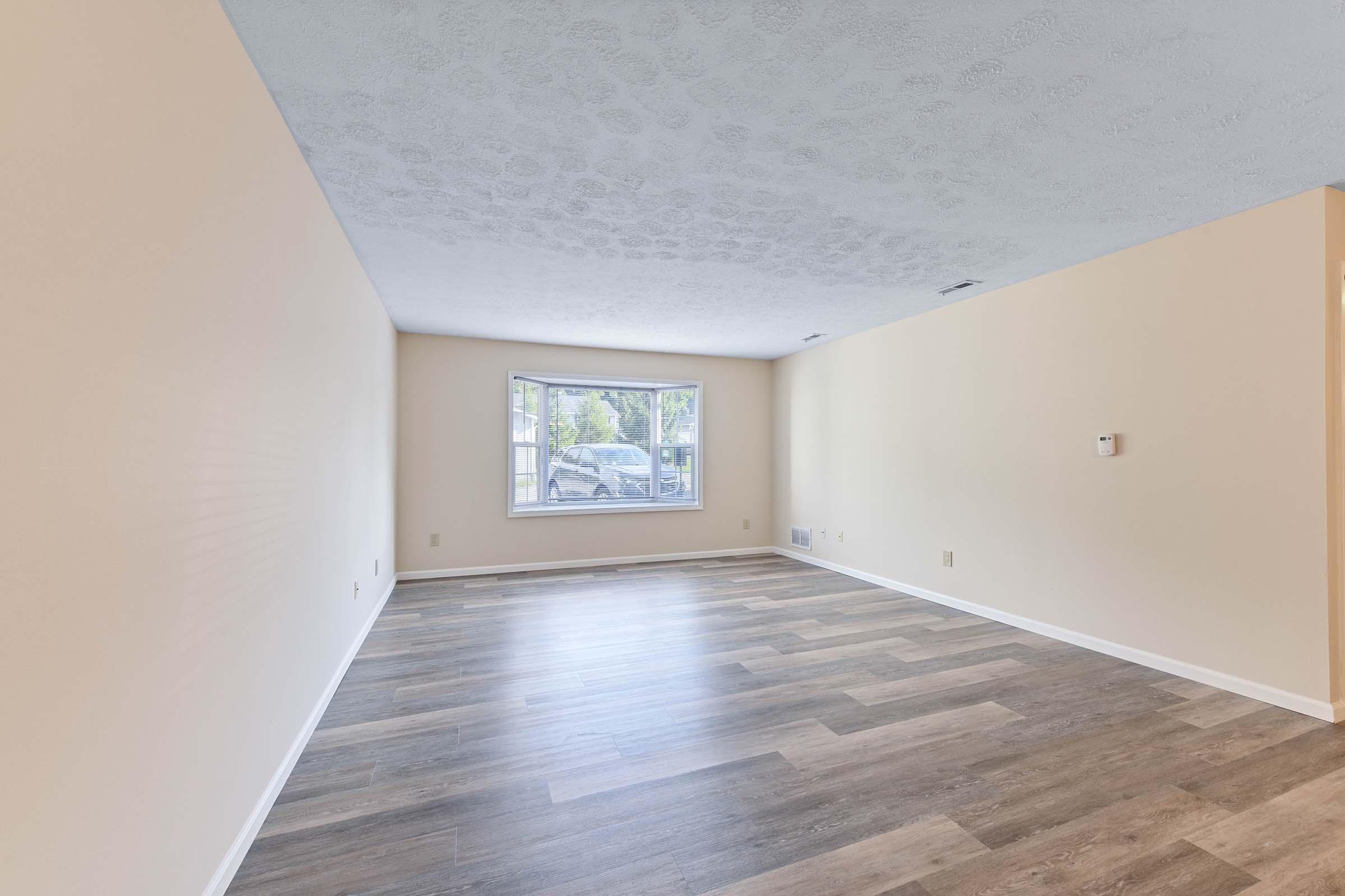 Empty room with light beige walls and a textured ceiling. The floor features brown laminate planks. A large window allows natural light to fill the space, showing greenery outside. The room has no furniture or decorations, emphasizing its spaciousness and potential for customization.