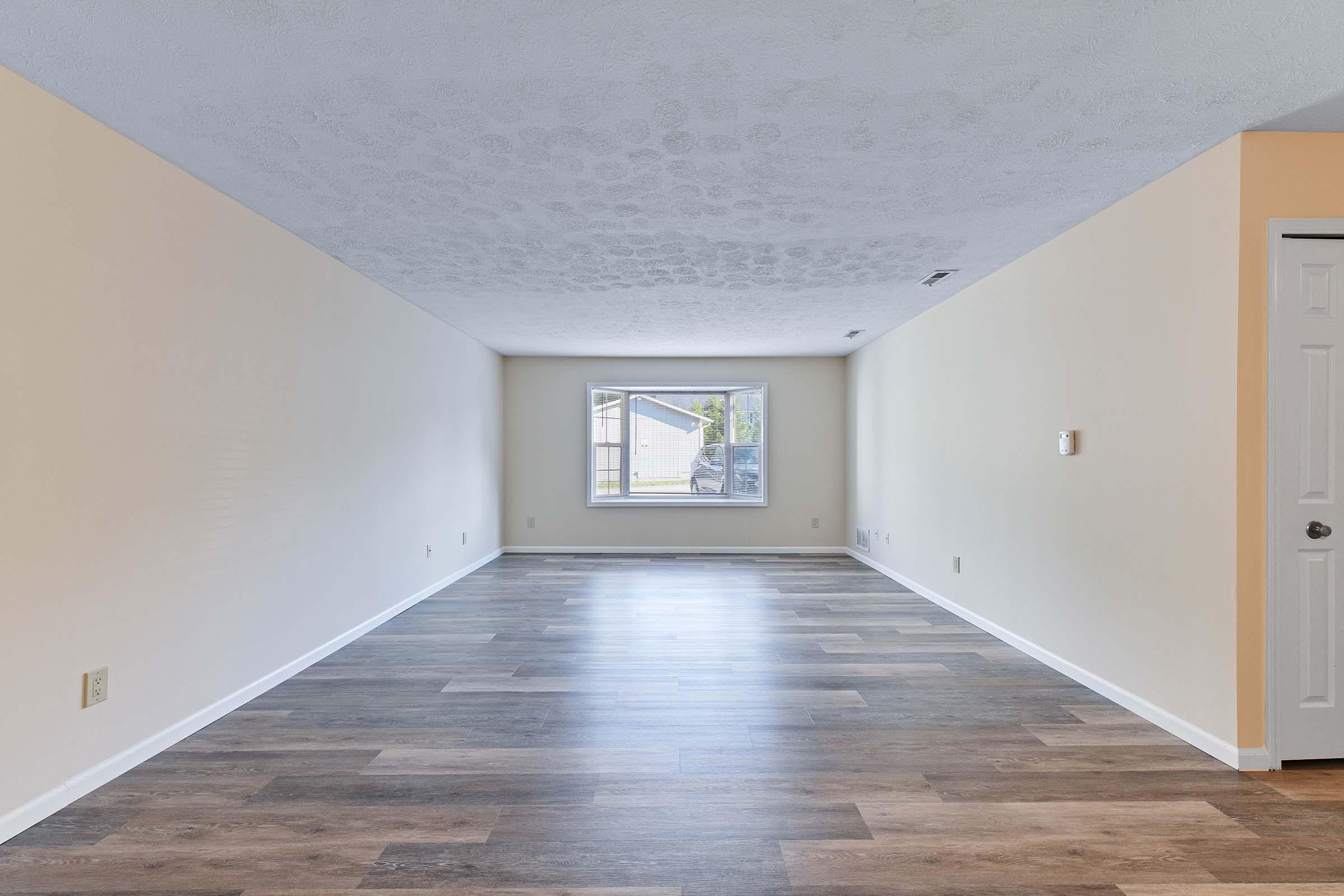 Spacious interior room with light-colored walls and a large window letting in natural light. The floor features a wood-like finish, offering a modern and clean appearance. The area is uncluttered, creating a versatile space for various uses.