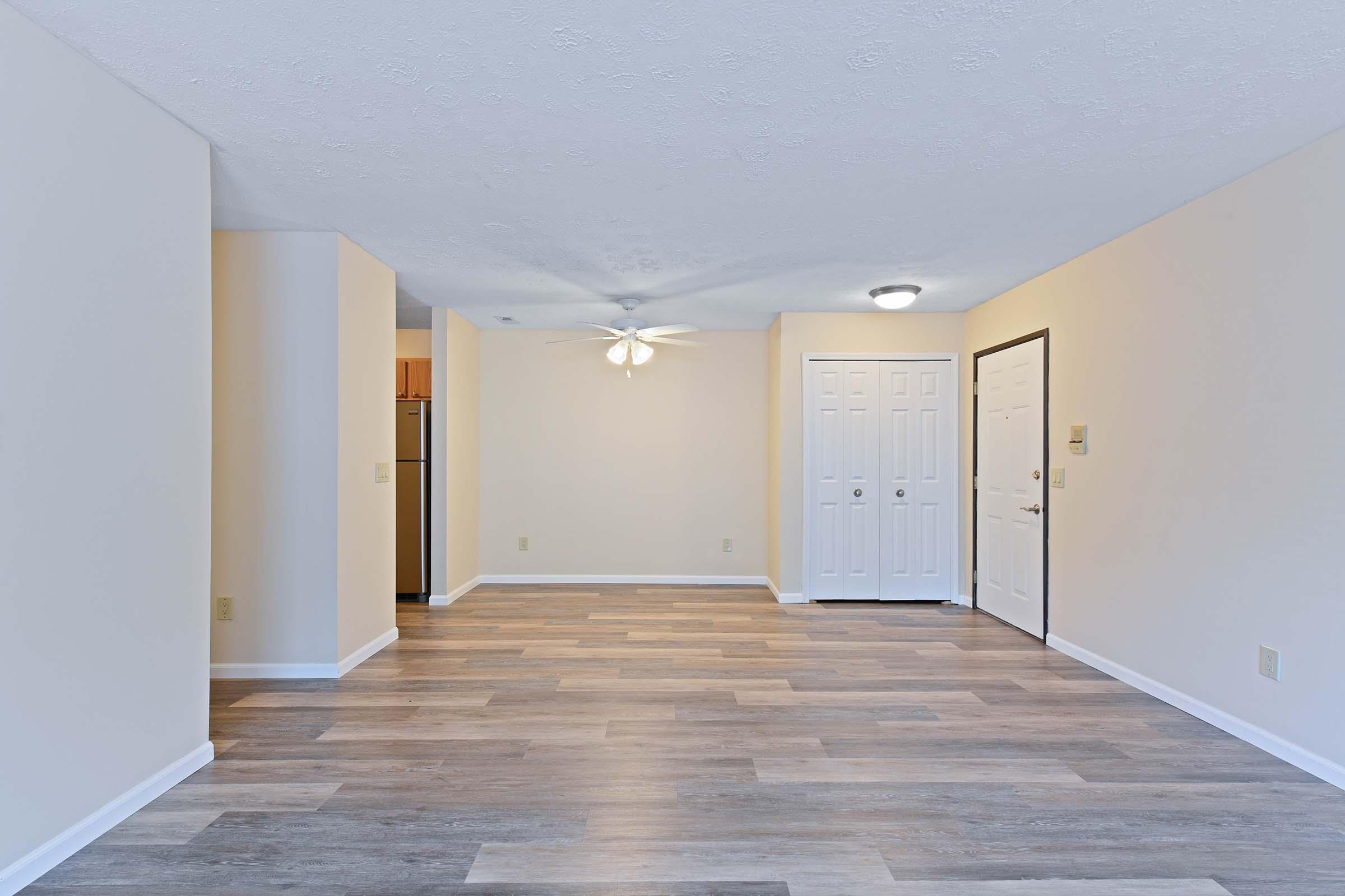 A spacious living area in a modern apartment featuring light-colored walls, vinyl flooring, and a ceiling fan. The room is open and airy, with a doorway leading to a kitchen area visible in the background. Two white doors are present, likely leading to different rooms, and ample natural light brightens the space.
