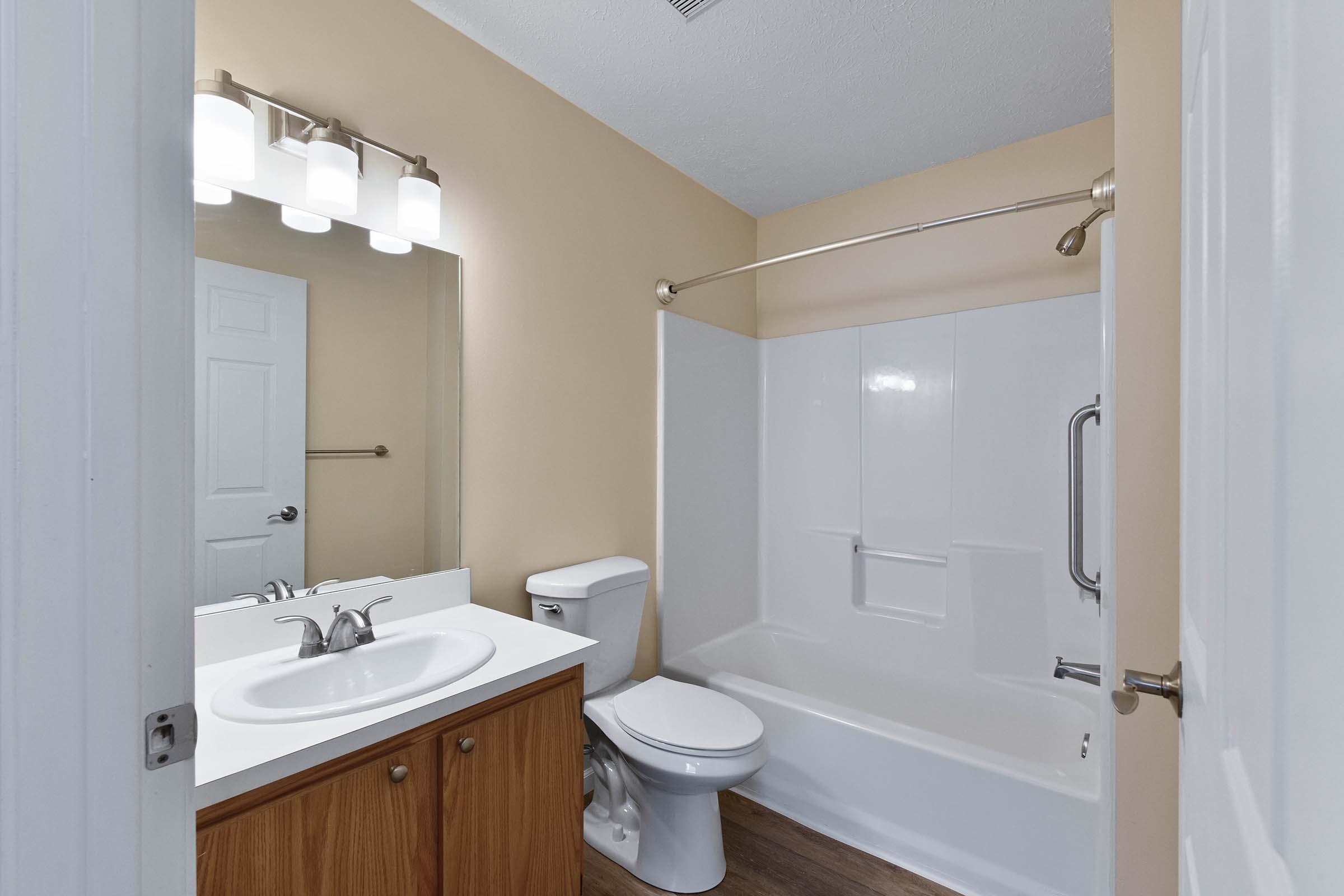A clean and modern bathroom featuring a white bathtub, toilet, and a sink with a wooden cabinet. The walls are painted a light beige, and there is a large mirror with overhead lighting above the sink. A towel bar is mounted on the wall, and the floor has a brown wood finish.