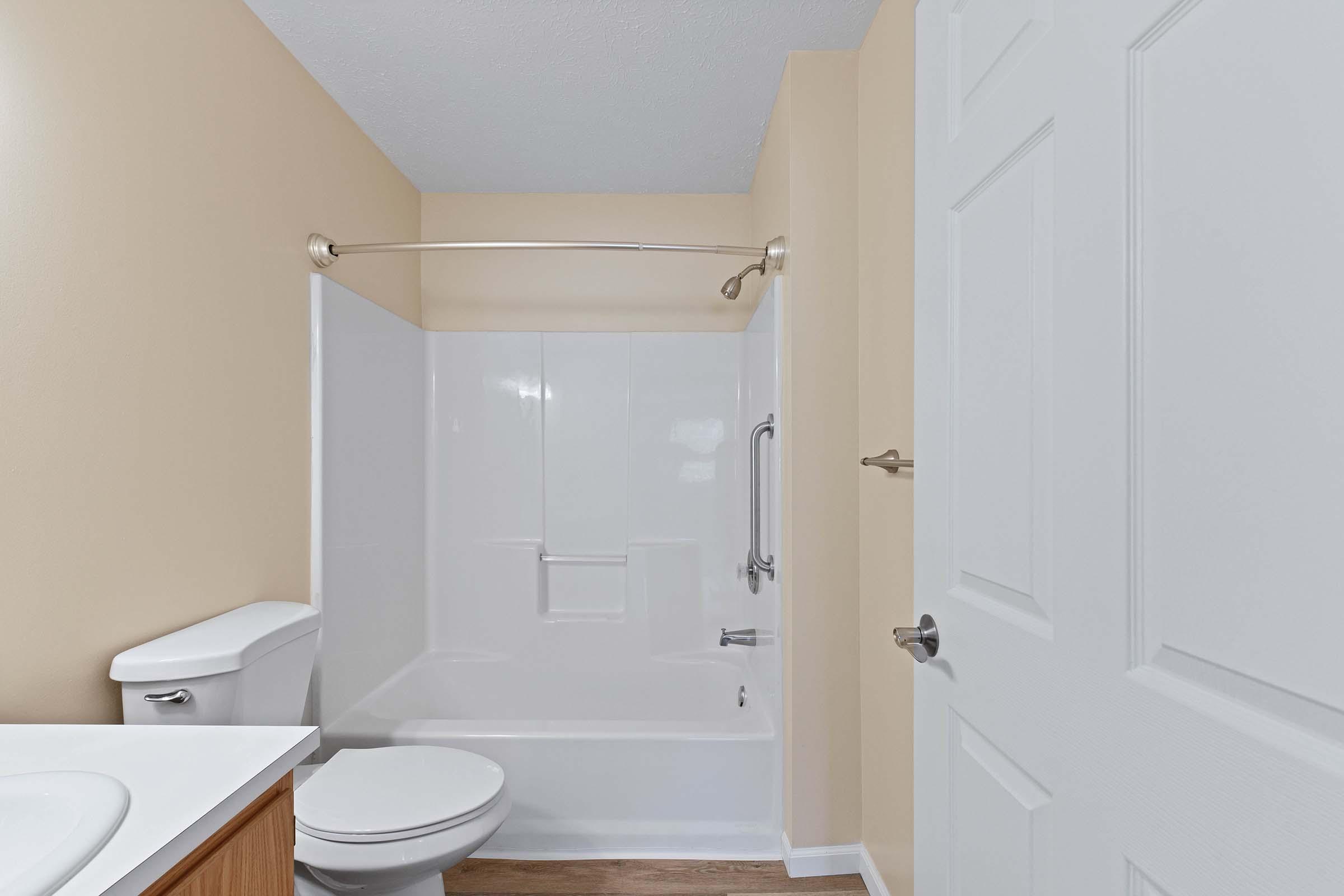 A clean and simple bathroom featuring a white bathtub with a shower curtain rod, a toilet, and a sink with a light wood cabinet. The walls are painted a soft beige color, and there is a white door visible in the background, contributing to a bright and spacious feel.