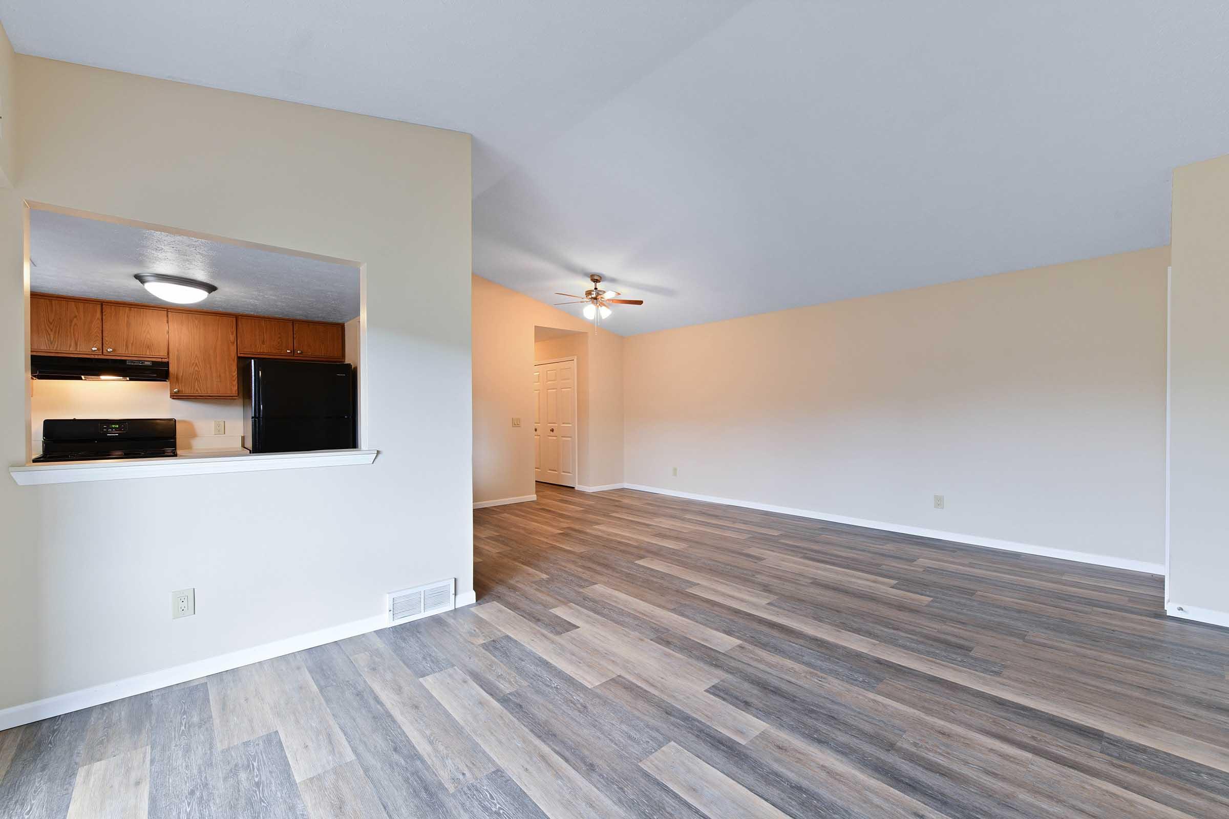 Spacious living area with wood-style flooring, featuring a view into a kitchen area with wooden cabinets and black appliances. The room is brightly lit with natural light and includes a ceiling fan. Walls are painted in a light color, creating an open and inviting atmosphere.