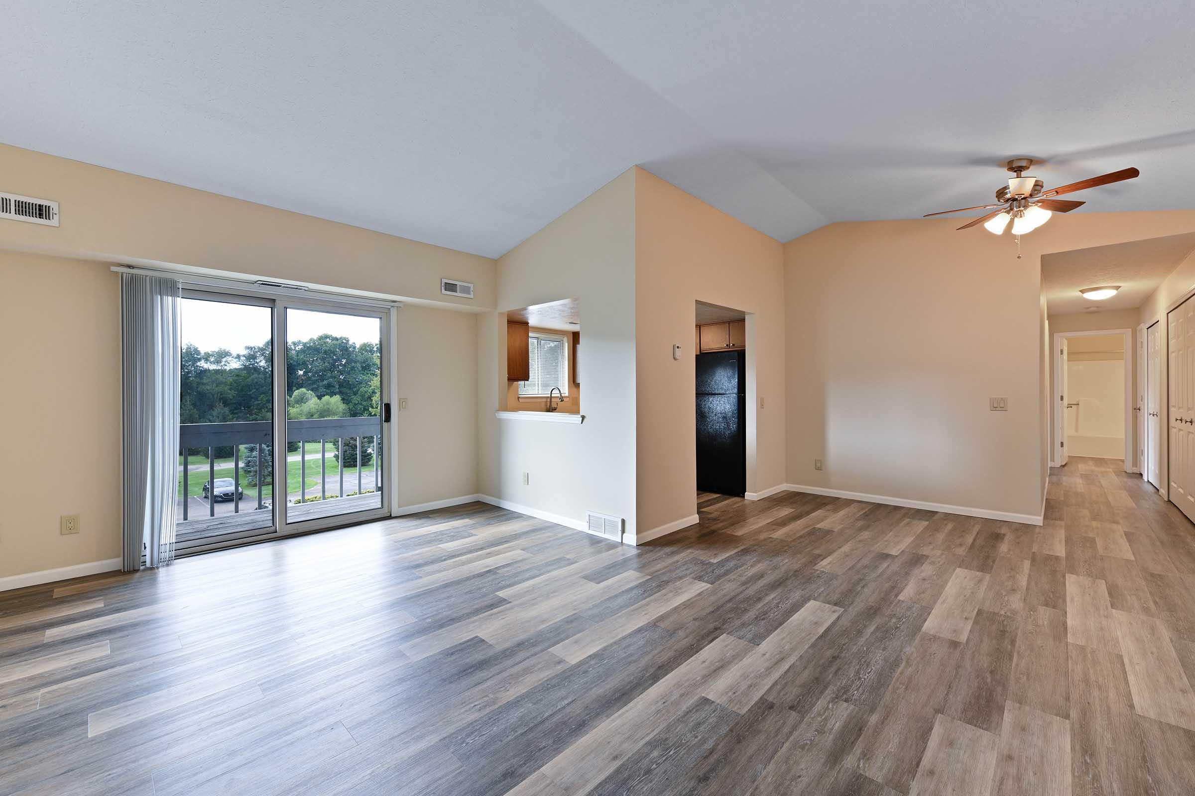 Spacious interior of an apartment featuring large windows, wooden flooring, and a ceiling fan. The layout includes a living area that opens to a balcony, a kitchen nook with a view, and a hallway leading to additional rooms. Natural light enhances the inviting atmosphere.