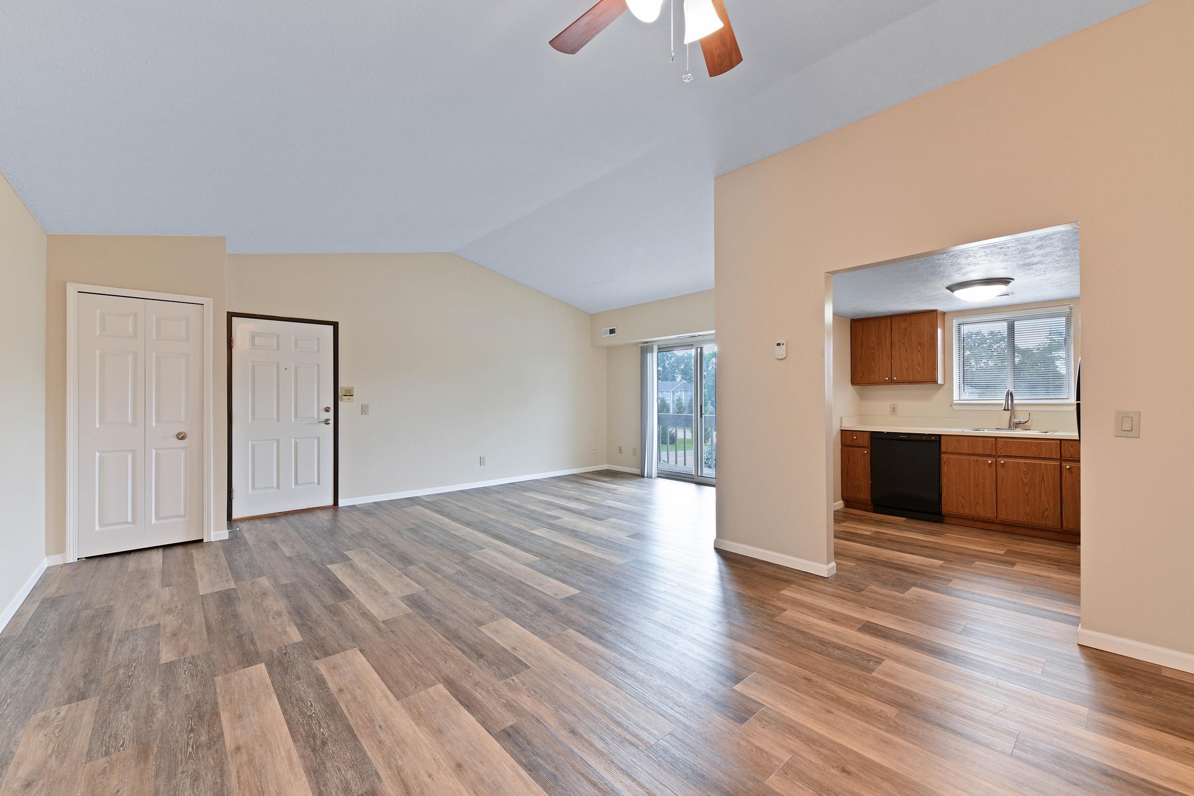 Spacious living area featuring wood-look flooring, vaulted ceiling with ceiling fan, and a large opening to a kitchen area with wooden cabinets. Two doors lead to the entrance, and sliding glass doors provide access to an outdoor space. The room is well-lit with natural light.