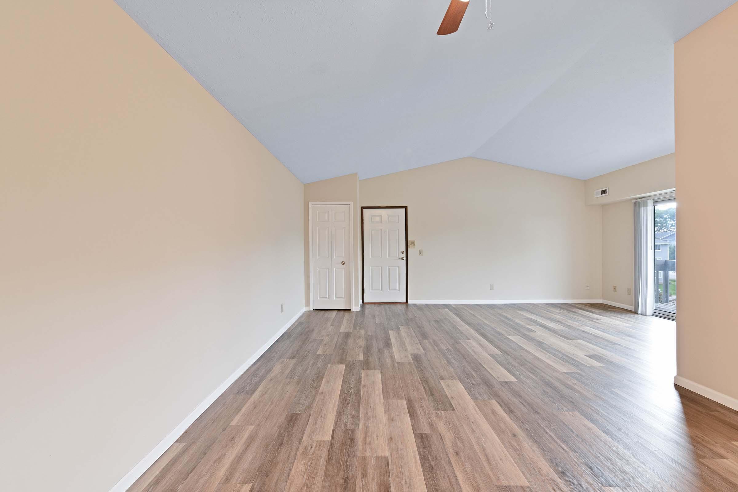 Empty interior of a spacious room with a high ceiling, featuring light-colored walls and wooden flooring. There are two doors on one wall, and a large sliding glass door leading to an outdoor area. The overall ambiance is bright and airy, suitable for various interior design styles.