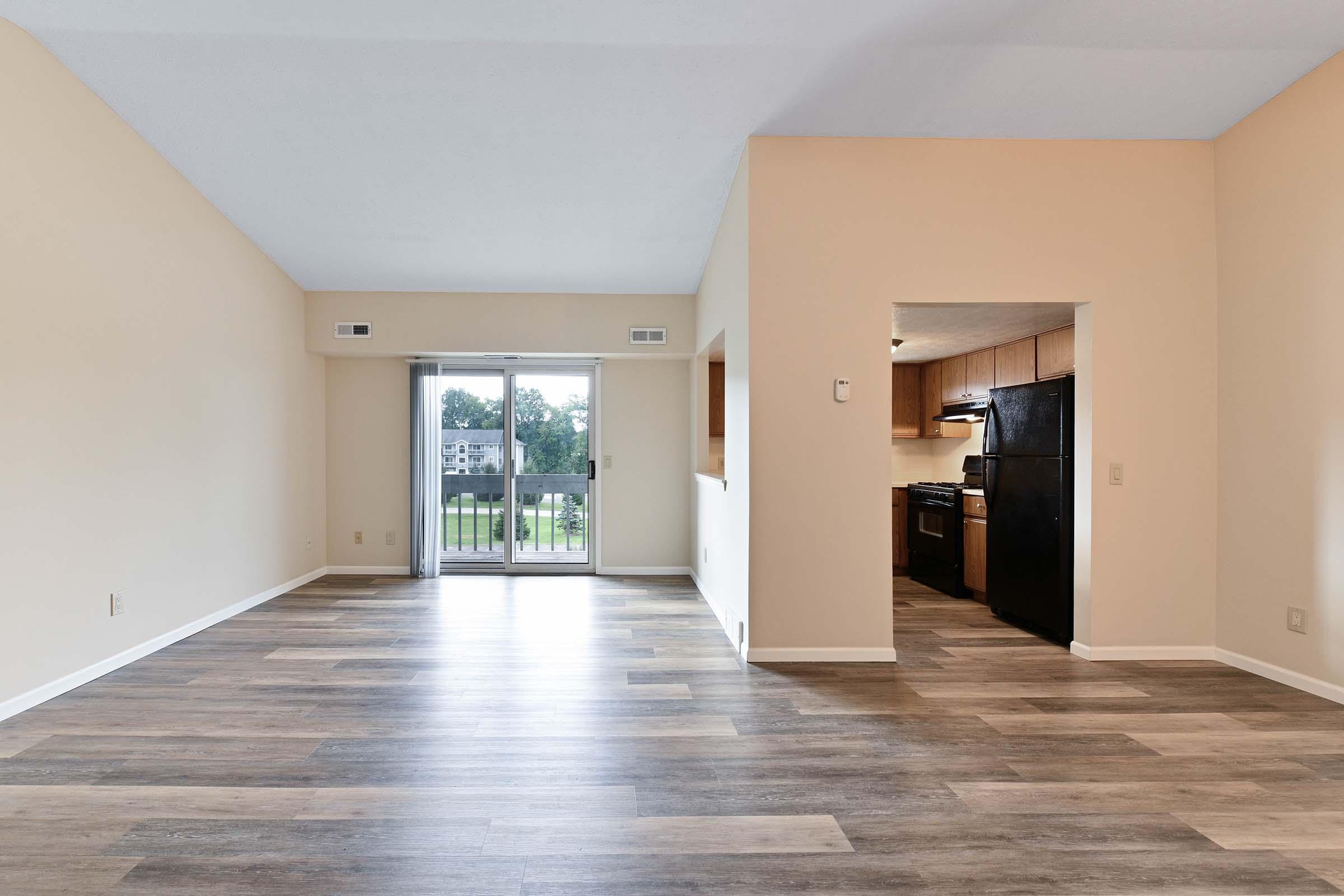 Spacious light-filled living room with neutral beige walls and wooden floors. A sliding glass door leads to an outdoor balcony, while a doorway on the right opens to a kitchen featuring black appliances. The open layout creates an inviting atmosphere.