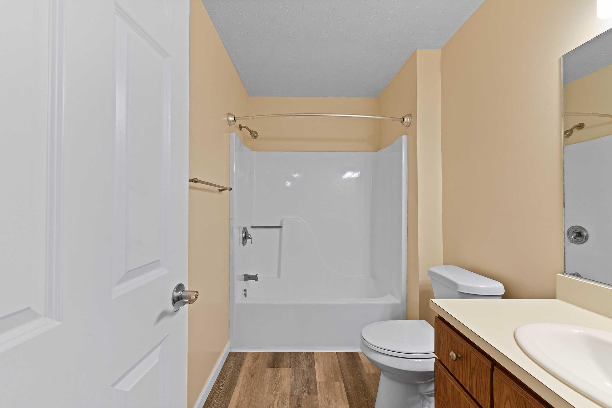 A clean and well-lit bathroom featuring a white bathtub with a shower curtain rod, a toilet, and a vanity with a sink. The walls are painted a warm beige, and the floor is covered in wooden laminate. A mirror is mounted above the sink, creating a bright and inviting atmosphere.