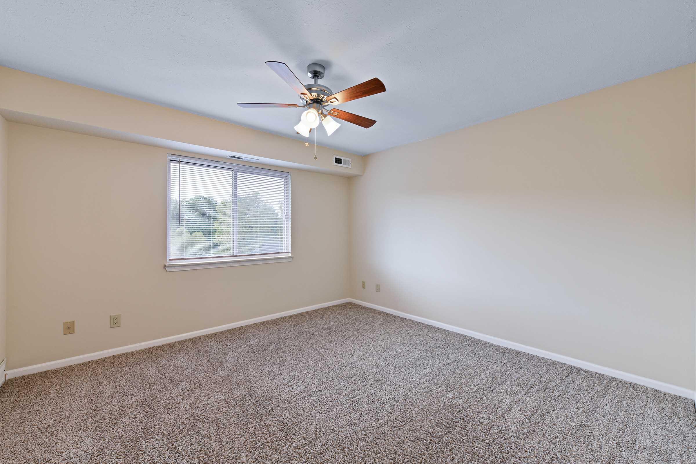 Spacious room with light beige walls and a ceiling fan featuring wooden blades. Natural light streams in from a window with blinds, and the floor is covered in soft, neutral carpet. The room is empty, highlighting its potential for various uses or furnishings.