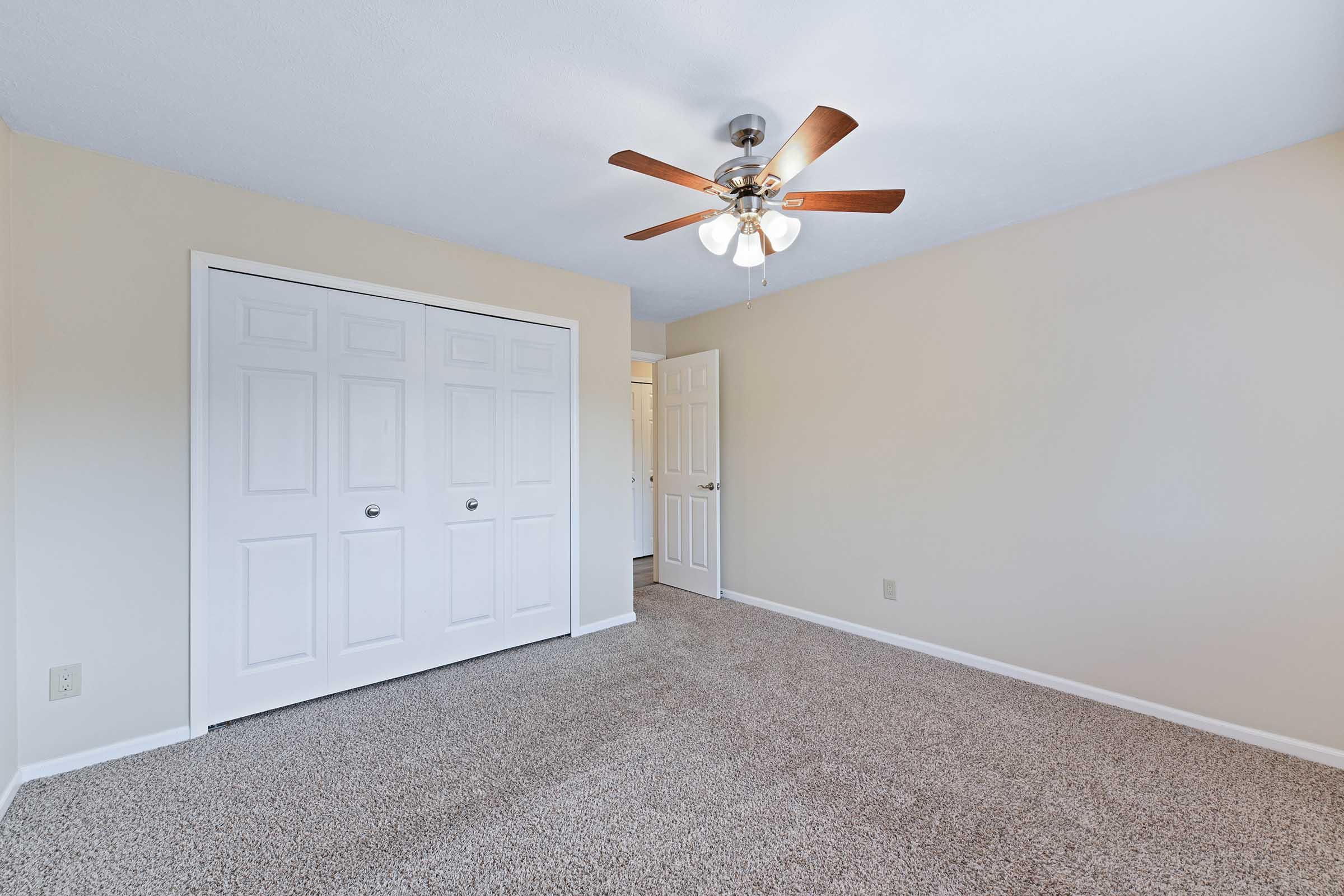 A spacious, well-lit bedroom featuring light beige walls, a ceiling fan with wooden blades, and plush carpet. There are two sets of white double doors on the left, providing access to storage or a closet. The room has a neutral and inviting atmosphere, suitable for various decor styles.