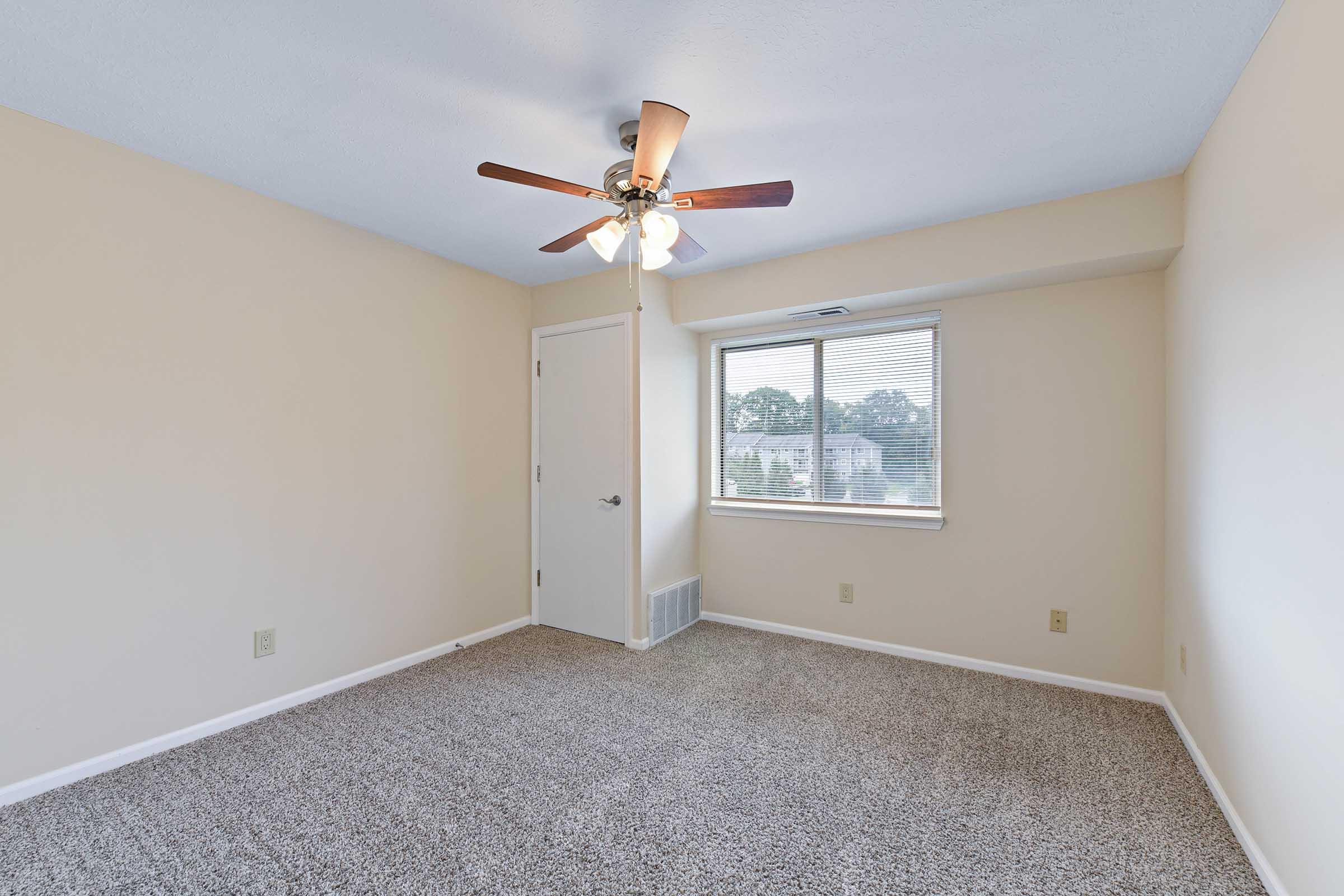 A well-lit, empty bedroom featuring beige walls and carpet. A ceiling fan with wooden blades hangs above, and a window provides natural light. A door leads to another room, and there are no furnishings, creating a spacious and clean atmosphere.