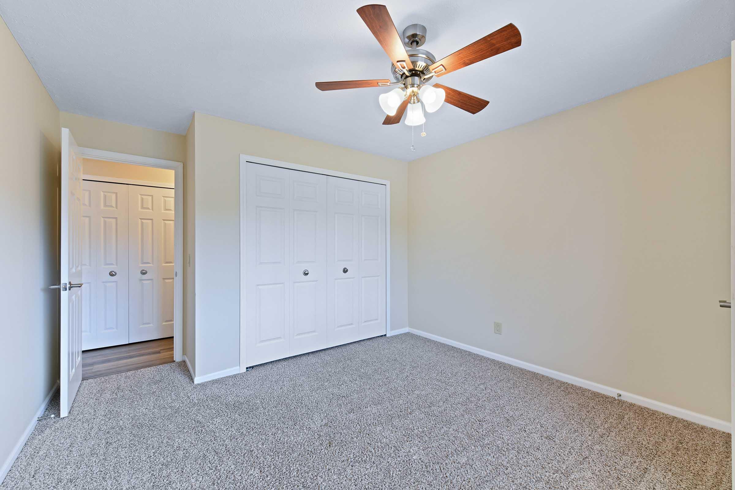 A well-lit empty bedroom with beige walls and carpeted flooring. It features a ceiling fan with light fixtures, double closet doors, and an open doorway leading to another room. The overall space is clean and airy, suitable for furnishing.
