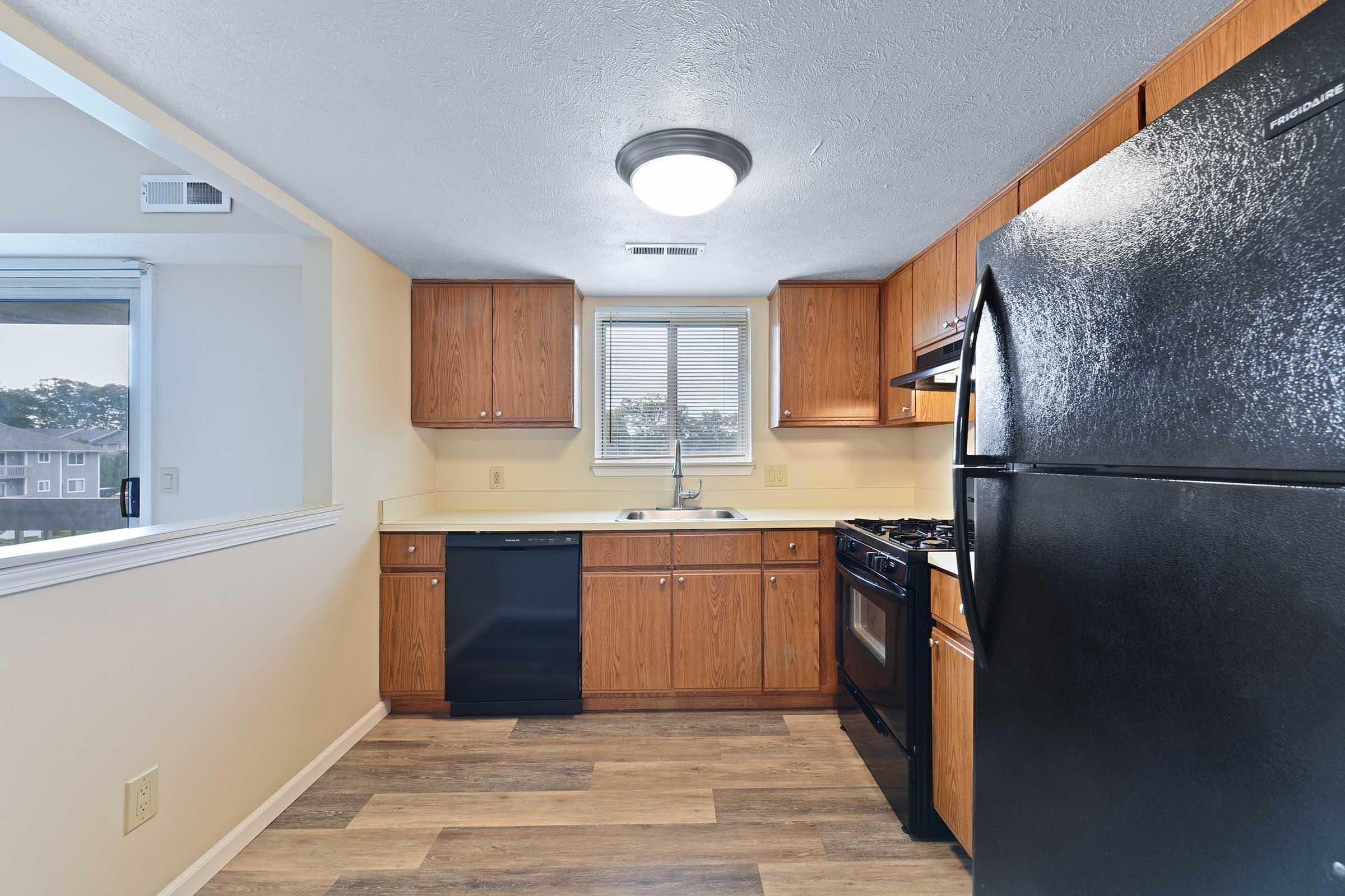 A kitchen featuring wooden cabinets, a black refrigerator, a black oven, and a dishwasher. The space has a window letting in natural light, with a neutral-colored wall and wood-like flooring. The overall design is modern and functional, suitable for cooking and meal preparation.