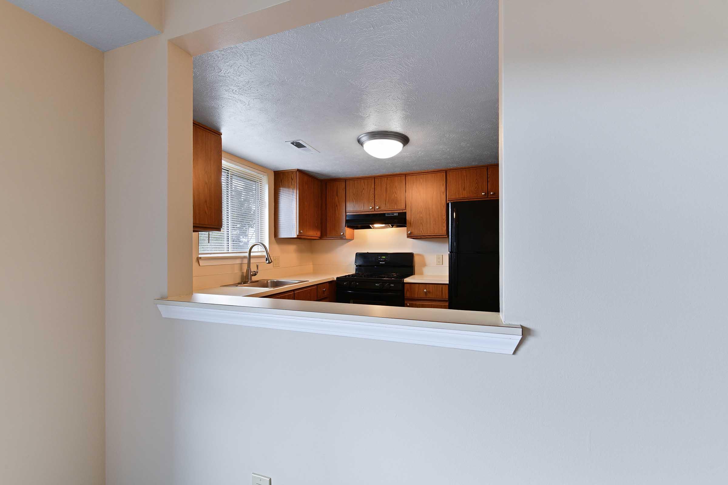 A view of a modern kitchen through a wall cutout. The kitchen features wooden cabinets, a black stove, and a refrigerator. There is a sink by a window allowing natural light, with overhead lighting illuminating the space. The walls are painted in a light color, creating a bright and inviting atmosphere.