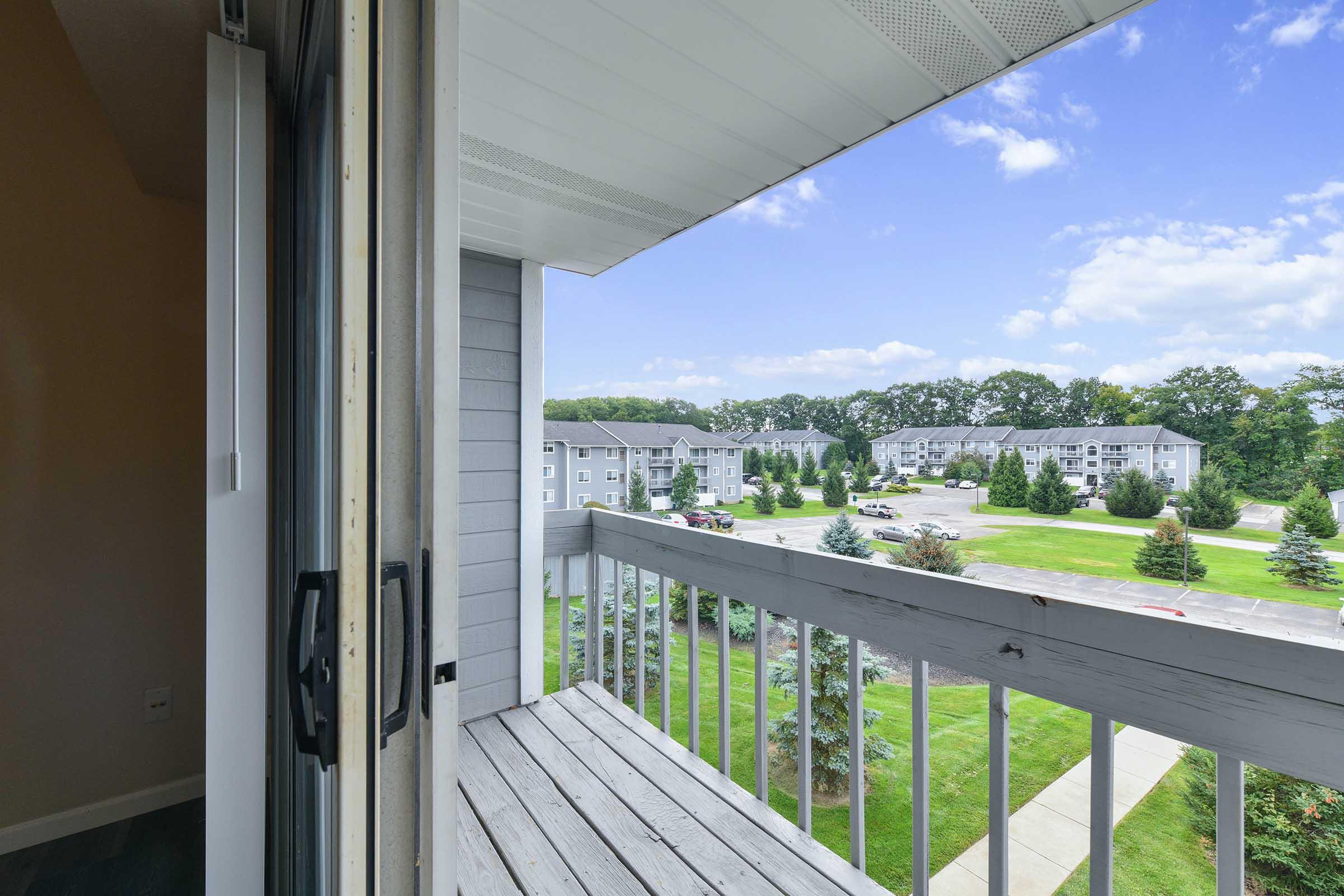 A view from a balcony overlooking a landscaped area with trees and grass. In the background, there are several apartment buildings. The sky is partly cloudy, creating a bright and inviting atmosphere.