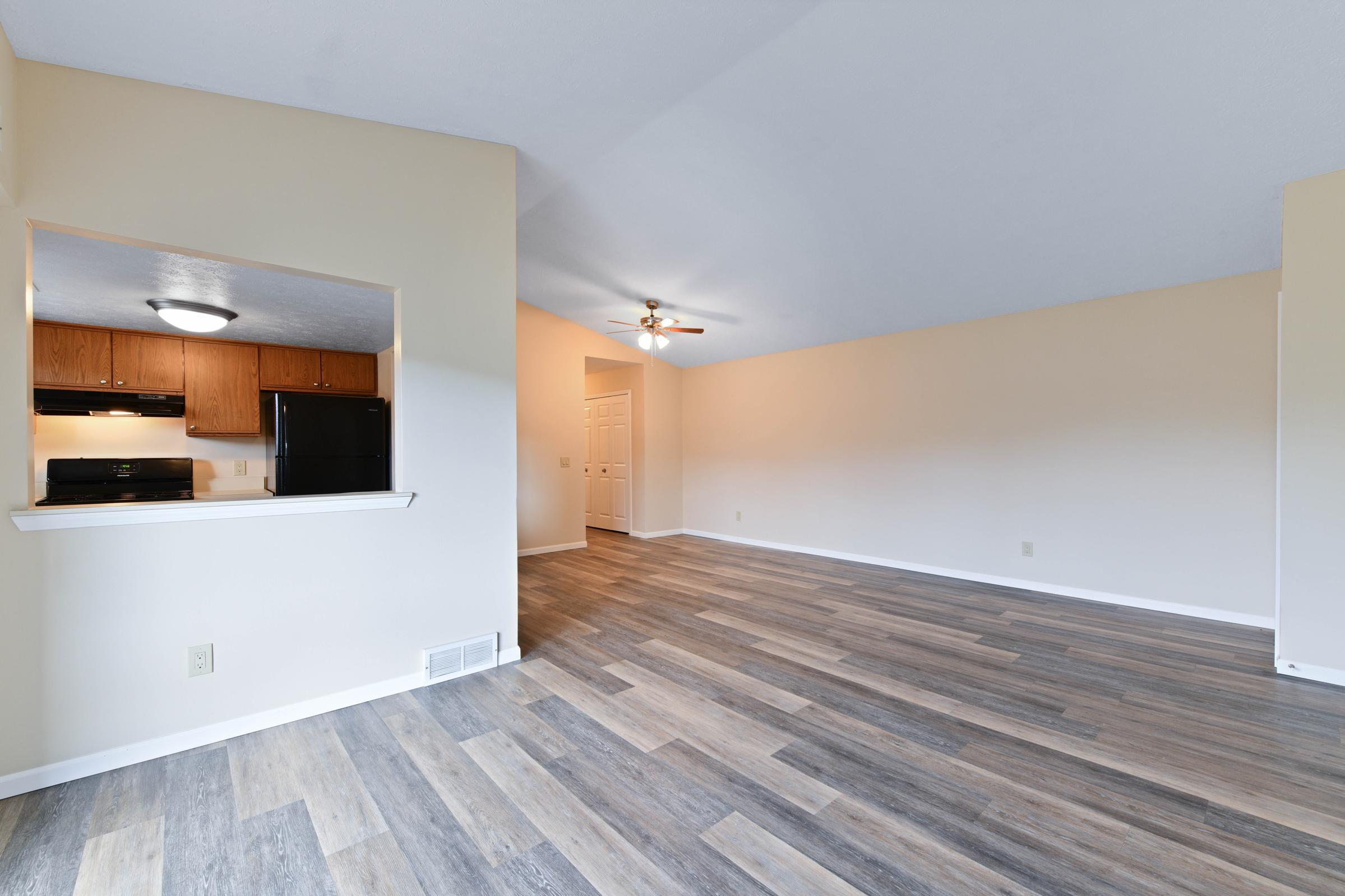 Spacious, well-lit living area featuring light hardwood-style flooring, a ceiling fan, and an open layout. A kitchen area is visible in the background with modern appliances. The room has neutral-colored walls, providing a blank canvas for personalization. Ideal for comfortable living and entertaining.